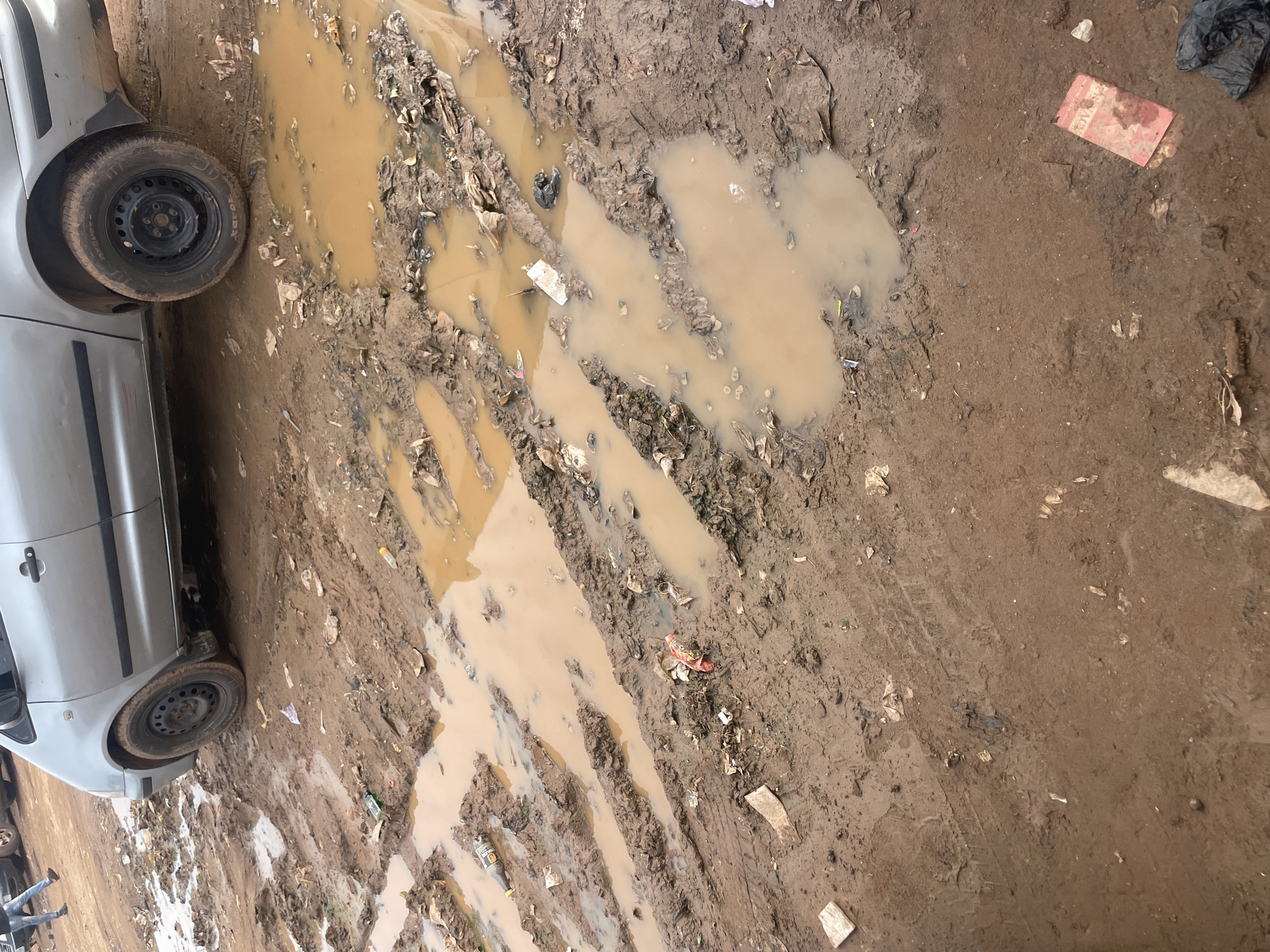 The scene depicts a deeply rutted, unpaved road or open area in Nyanya, Nigeria, characterized by extensive mud and water accumulation. A silver four-door sedan is positioned on the right side of the frame, with both visible wheels (front and rear) heavily soiled with brown mud, indicating travel through the challenging terrain. The ground is saturated, forming numerous shallow to moderately deep puddles of turbid, light brown water within prominent vehicle tire tracks that traverse the area. Scattered throughout the mud and within the puddles is various discarded refuse, including several plastic bottles (one dark brown, one clear), fragments of white and black plastic bags, pieces of paper, and other unidentifiable debris. In the lower right foreground, a red, rectangular object, possibly a book or package, is partially submerged in a puddle, adjacent to a black plastic bag filled with trash. In the upper left background, a person wearing blue jeans and dark shoes is partially visible, walking on a less muddy section of ground. The overall environmental condition suggests poor drainage and recent precipitation.