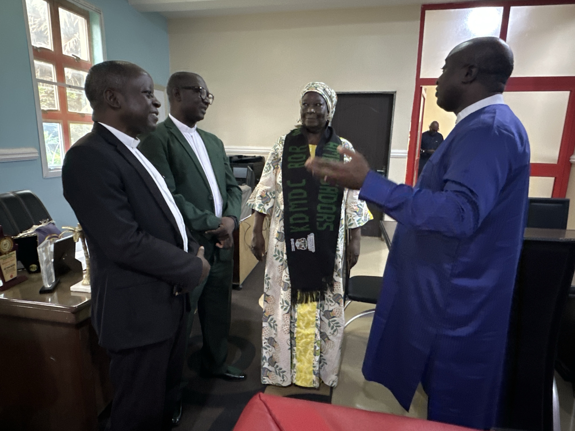 This media file depicts four people indoors, seemingly engaged in a formal discussion or meeting in what appears to be an office or meeting room in Goni Gara, Nigeria.
On the left, a man in a black suit and white shirt is partially visible, facing towards the right. To his right stands another man wearing a green suit jacket over a white shirt and collar. Next to him is a woman wearing a colorful patterned dress with a distinctive black scarf around her neck. The scarf has green lettering on it, which appears to read "KDMDCROR" and "ADOR" possibly indicating an organization or affiliation. She is looking towards the man in the blue garment. On the far right, a man in a vibrant blue traditional outfit is facing the woman, gesturing with his right hand as if speaking. In the background, another man in a dark blue outfit is visible through an open doorway.
The scene is indoors, with muted lighting and the presence of office furniture like desks and chairs. A window with a red grid pattern is visible on the left, and part of a red chair is in the foreground. The time of day cannot be definitively determined from the image. The overall mood appears to be one of engagement and discussion.