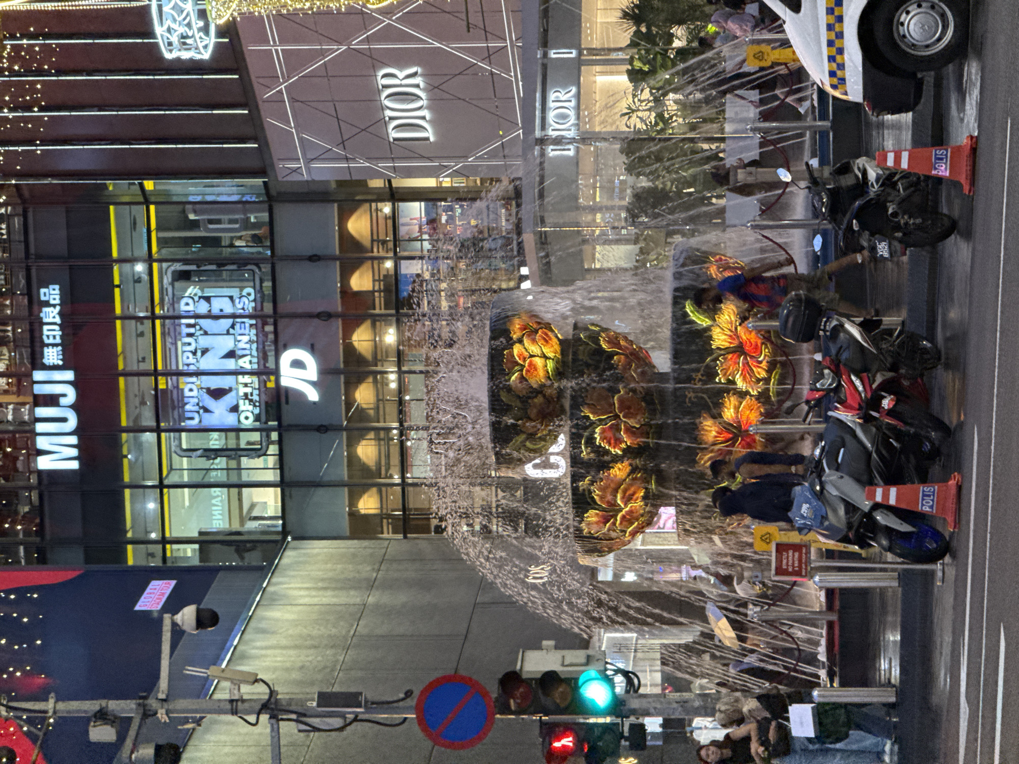 The image shows a nighttime street scene in Kuala Lumpur, Malaysia, featuring a fountain in the foreground. The backdrop includes several prominent retail stores, including MUJI, JD Sports, and Dior, each displaying large signage. Pedestrians are visible on the street near the fountain. Two motorcycles are parked partially on the sidewalk near traffic control devices such as a police cone, traffic lights displaying both red and green, and a no-parking sign. A police car is parked close by. The fountain itself is adorned with floral decorations and illuminated by lights. The overall ambiance is urban, with modern architecture and commercial activity.