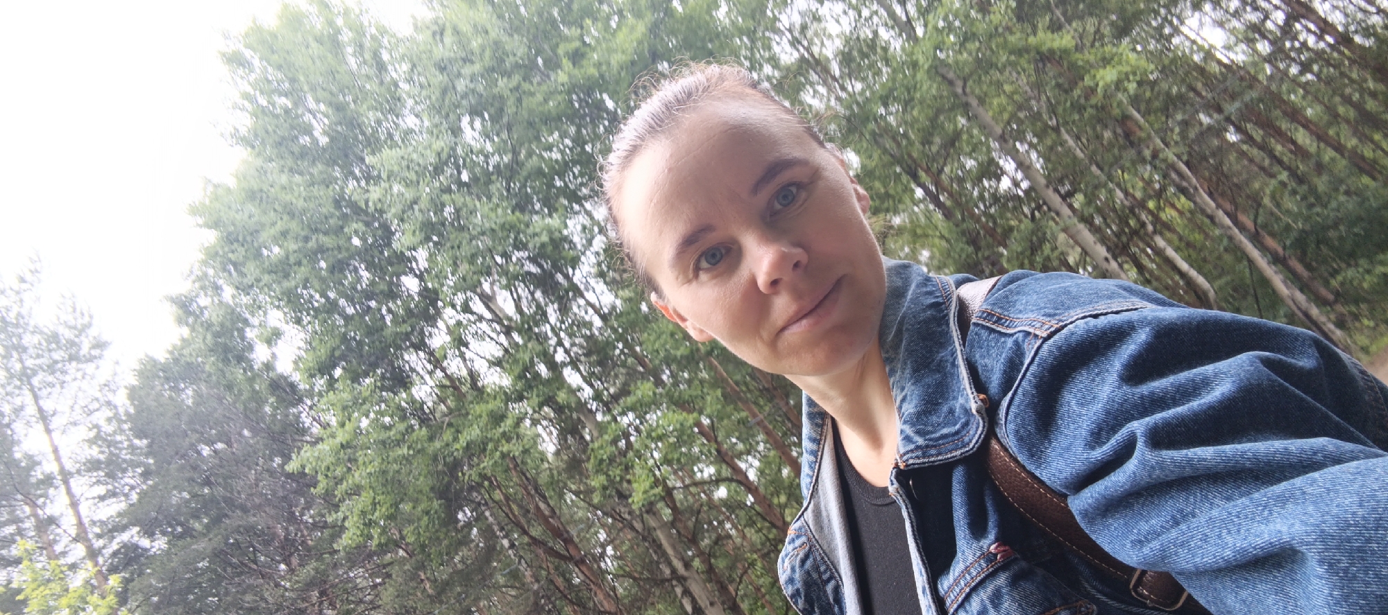A woman is depicted in a low-angle, upward-facing self-portrait, positioned in the lower-right quadrant of the frame. She has light brown hair pulled back, blue eyes, and fair skin, maintaining direct eye contact with the camera with a neutral expression and a slight smile. She is wearing a blue denim jacket with visible stitching and a collar, over a dark (possibly black) top. A brown strap, consistent with a shoulder bag, is visible over her right shoulder. The background consists of a dense forest environment, typical of the Chaikovsky, Russia area. Tall, slender trees with light-colored, possibly birch-like trunks and green deciduous foliage dominate the right and center, extending diagonally upwards across the frame. On the far left, taller coniferous trees with darker green needles are visible. The upper left portion of the image shows an overcast, bright white sky, indicating diffuse natural light.