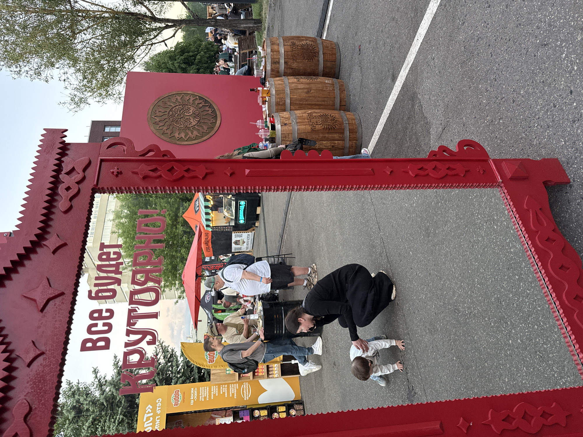A ground-level medium shot captures a lively outdoor marketplace setting, seemingly in Moscow, Russia, as inferred from the Russian text on a prominent red sign that reads "Все будет КРУТОЯРНОЕ," meaning "Everything will be cool, steep, and bright." The sign, designed with a traditional, carved aesthetic, acts as a partial frame for the scene.
The foreground features a woman in a black outfit crouched down near a young child, who is on their hands and knees on the asphalt. The interaction between the woman and child is centered in the frame.
Behind them, various market stalls are visible, showcasing an array of products and food vendors. Several people are milling around these stalls, suggesting a bustling event. In the background, three wooden barrels are placed next to a tall red wall adorned with a circular design. A person is lying down next to the barrels, adding to the casual, open-air atmosphere. In the far background, there is a building and several people sitting at tables and chairs.
The overall impression is that of a vibrant, community event in a public space. The sky is visible between buildings and trees.