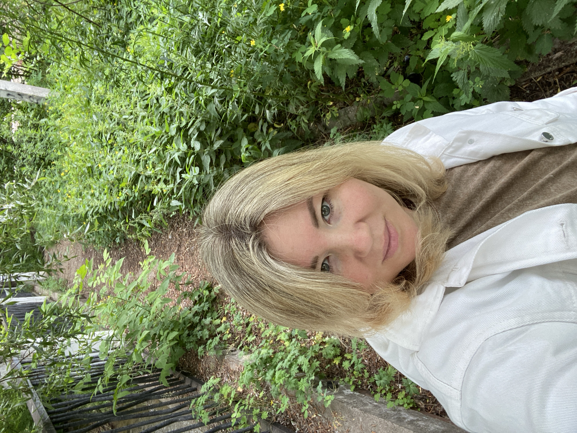 This is a slightly angled, medium shot of a woman taking a selfie outdoors. She has shoulder-length blonde hair and is wearing a white denim jacket over a brown t-shirt. The background is filled with lush green foliage, including plants with serrated leaves and small yellow flowers, suggesting a natural or park-like setting. The lighting indicates it is daytime, possibly late morning or afternoon, with soft, diffused light. The woman's expression is neutral, and the overall mood is casual and relaxed. The image appears to have been taken in Chaikovsky, Russia. There is no visible text in the image.