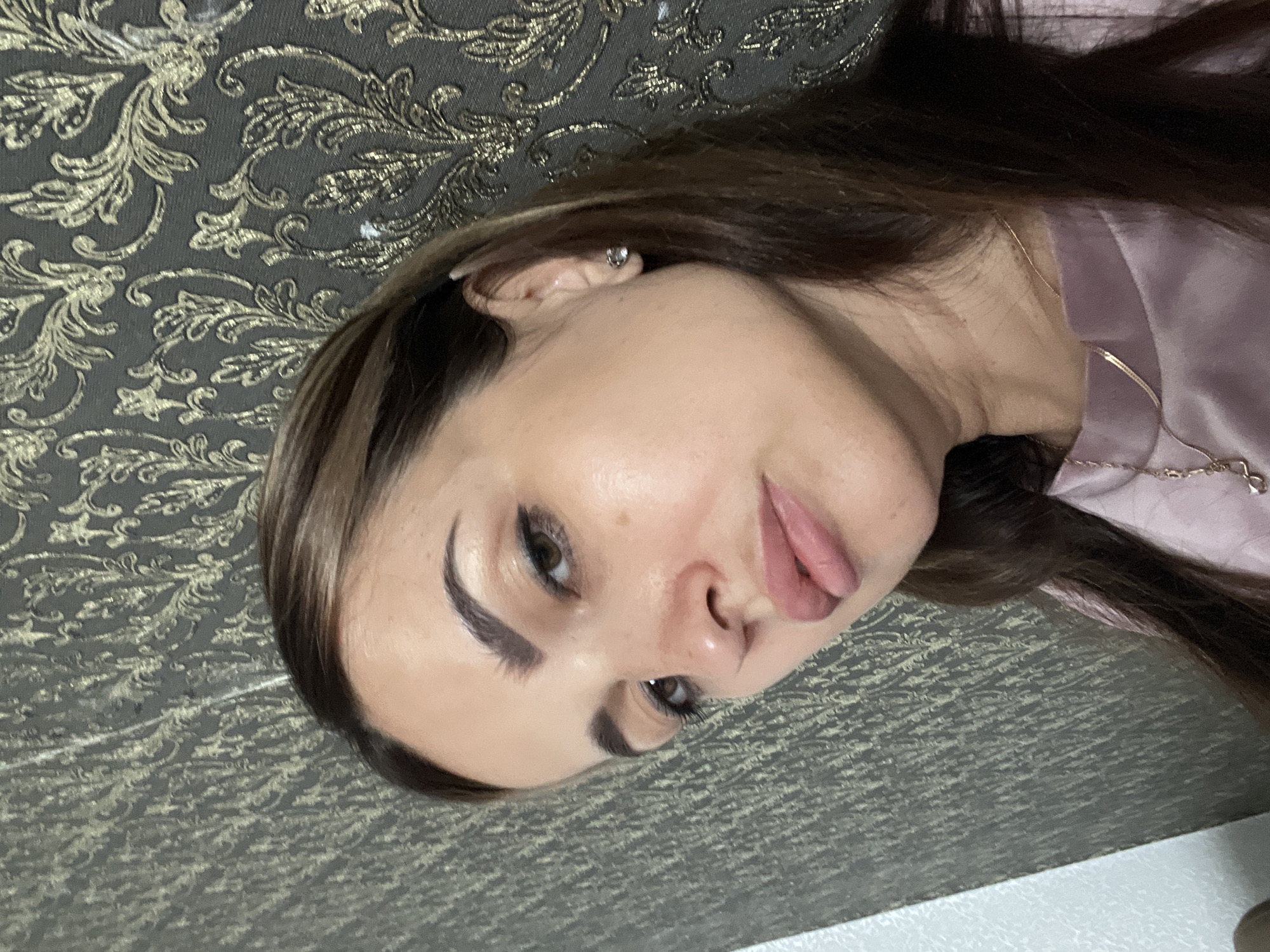 A close-up, head-and-shoulders shot captures a woman with long, dark brown hair and light skin. She is wearing a pink top and a delicate gold necklace with a pendant. Her right ear is adorned with a small, sparkling stud earring. The background is a textured wallpaper with a dark gray base and a repeating pattern of golden floral motifs. Her face is turned slightly to her left, with her gaze directed away from the camera. Her makeup includes defined eyebrows and eyeliner, with a subtle pink lipstick on her lips.