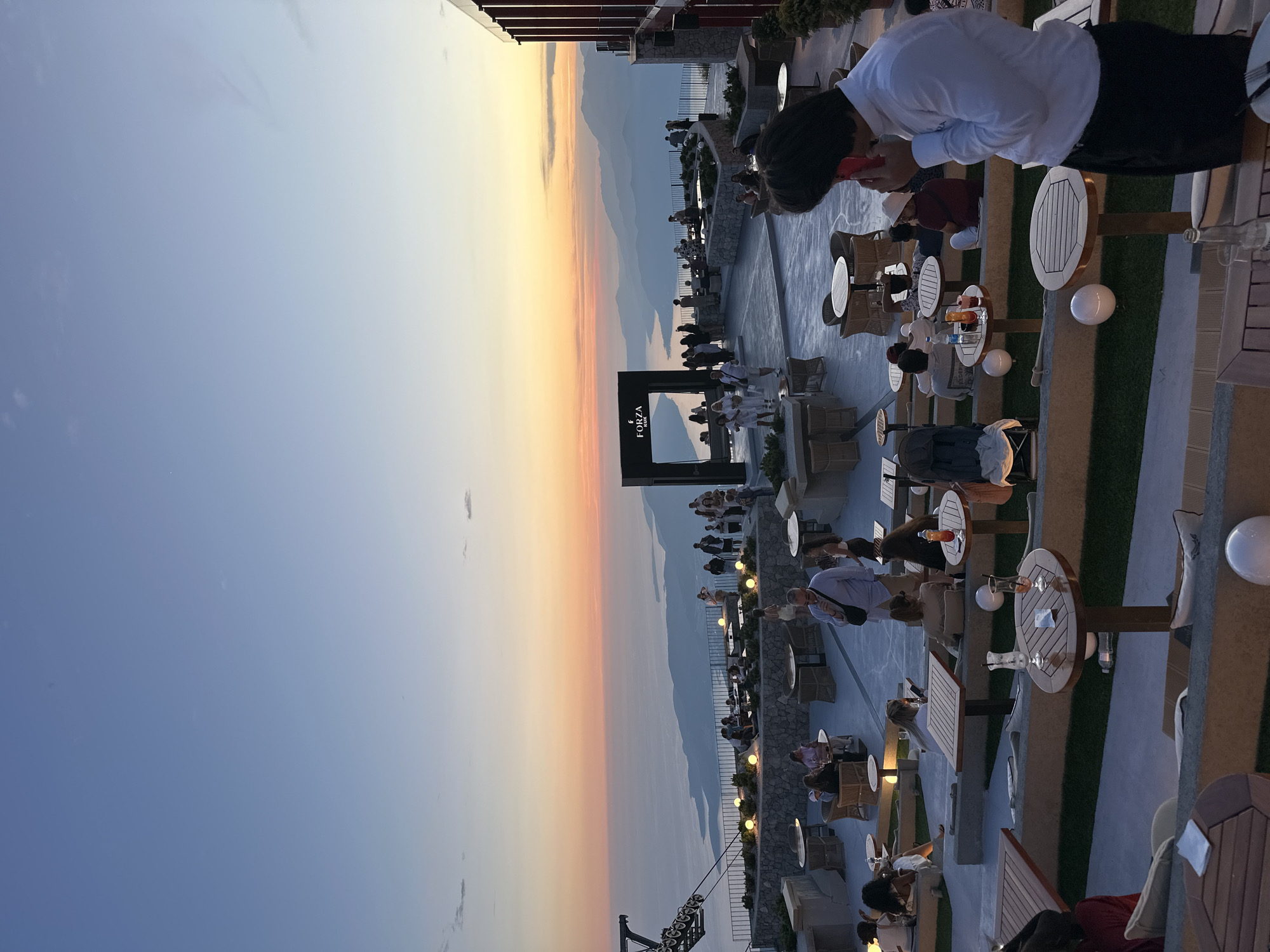 The image depicts an outdoor terrace overlooking a scenic view of the sea and mountains during sunset. The setting appears to be a restaurant or event venue, likely located in or near Kuk, Montenegro, given the coastal and mountainous landscape.
In the foreground, several tables and seating arrangements are visible, some occupied by people. To the right, a man in a white long-sleeved shirt and dark pants is on his phone, facing away from the camera. In the middle ground, more tables and patrons are seated, some dining and others conversing. A black archway with the "FORZA RUM" logo is present, suggesting a sponsored event or establishment. Beyond the terrace, the sea stretches out, with islands or peninsulas visible in the distance. The sky is painted with the warm hues of a sunset, transitioning from orange and pink near the horizon to a soft blue above.
In the background, a mountainous coastline is visible, with layers of hills receding into the distance. The architecture of the building on the right suggests a modern design, with balconies and dark facade elements. A cable car structure is also visible on the left side of the image, hinting at the elevated location of the venue. The overall ambiance is relaxed and sophisticated, with people enjoying the evening view and company.