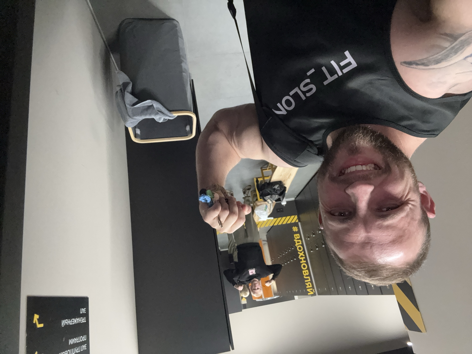 The image shows a smiling man taking a selfie within what appears to be a gym or fitness center. He is in the foreground, looking directly at the camera with a broad smile, revealing his teeth. He has short brown hair and a beard, and displays a muscular physique. He is wearing a black tank top with "FIT_SLON" written in white capital letters across the chest. A tattoo is visible on his left shoulder/upper arm. In his right hand, he holds a small bundle of blue and grey cloths, possibly gloves, along with a small metal object resembling a drill bit.
In the background on the left, an ironing board or similar flat surface with a grey cloth draped over it is visible. To the right and further back, another person is seen, possibly posing or flexing, wearing a black long-sleeved top and light-colored pants. Behind the main subject, there is a dark wall featuring metal lockers or storage units, decorated with yellow and black striped markings. On this wall, Russian text, including "#ВДОХНОВЛЯЙ" (Vdokhnovlyay, meaning "Inspire"), is partially visible. A sign in the lower left, when viewed upright, displays Russian text indicating different areas: "ЗАЛ ТРЕНАЖЕРНЫЙ" (Gym Hall) and "ЗАЛ ГРУППОВЫХ ПРОГРАММ" (Group Program Hall). The setting is indoors, well-lit, and suggests an active, fitness-oriented environment.
