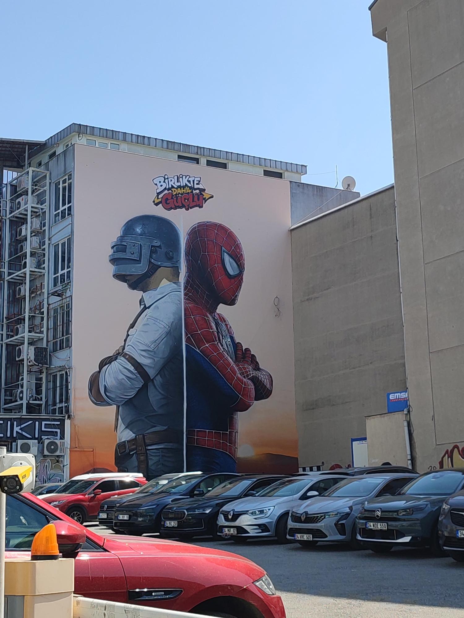 The image depicts a mural on the side of a building in Kadıköy, Turkey. The mural features two figures, one a character from the video game PUBG wearing a helmet and tactical gear, and the other is the superhero Spider-Man. They are positioned back-to-back, facing away from each other. The mural has text above the figures that reads "BİRLİKTE DAHA GÜÇLÜ," which translates to "Stronger Together."
In the foreground, a row of cars is parked. The cars include a red one in the closest foreground, followed by several other vehicles. The background shows the urban environment with buildings and a clear sky. The overall scene appears to be an outdoor area in a city, likely a street or parking lot, adjacent to a building with a large mural.