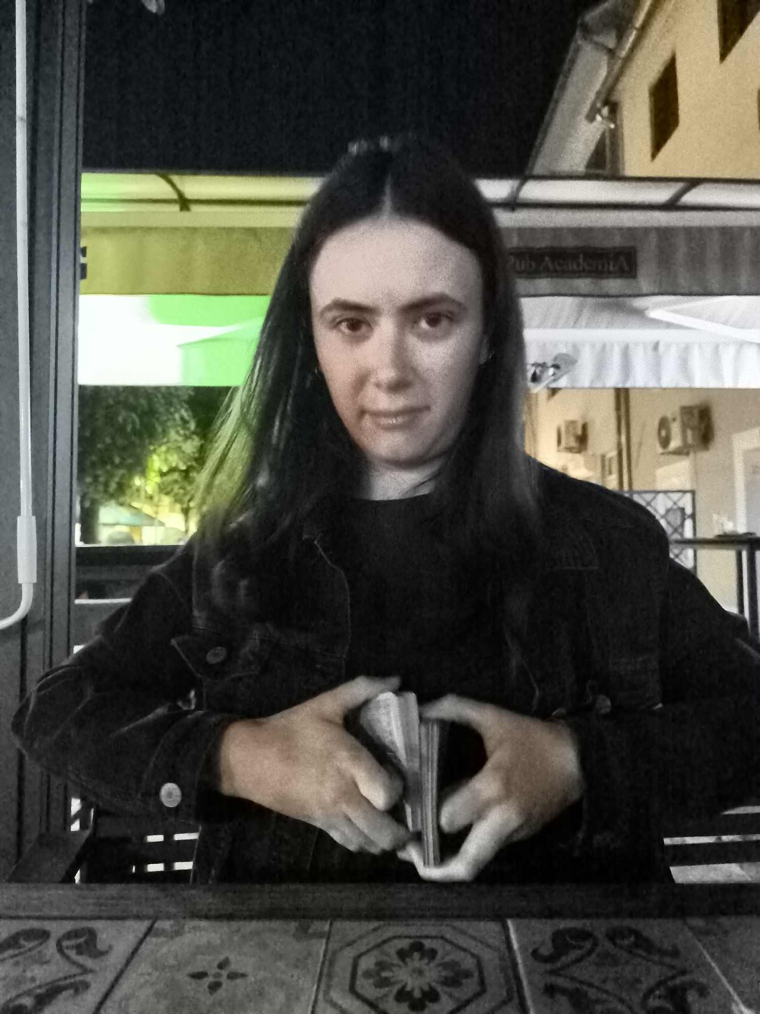 A person with long, dark hair and a dark denim jacket sits at a table, holding a deck of cards in their hands. The person's face is partially obscured by their hair, and they are looking towards the camera with a neutral expression. The background appears to be an outdoor seating area, possibly a cafe or restaurant, with a sign that reads "Pub Academia". The lighting is dim, suggesting it is nighttime or evening. The table in front of the person has a patterned surface. The overall scene is dimly lit, with a green glow visible in the background from some artificial lighting.