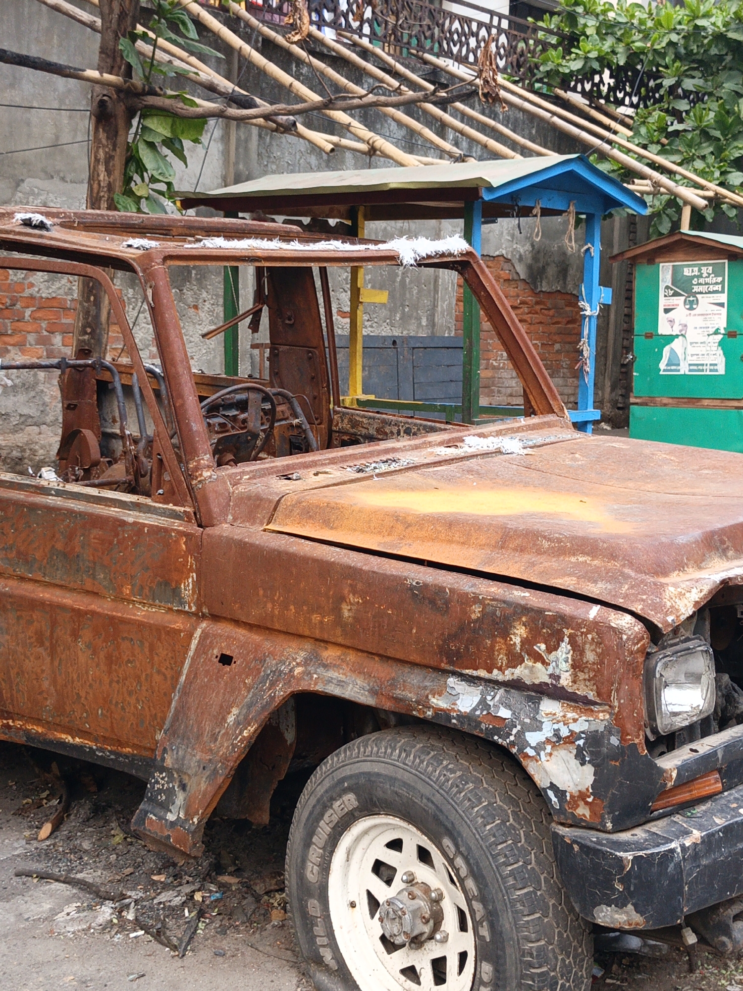 A heavily rusted, derelict vehicle with a white tire is parked in what appears to be an urban setting. The vehicle is missing its roof and doors, exposing the interior. The overall state suggests long-term neglect and weathering. The tire has the words "WILD CRUISER" written on it. A small blue structure and a green kiosk with a poster are visible in the background. Bamboo structures overhang the scene. Based on the signage and style of the environment, this is likely Dhaka, Bangladesh.