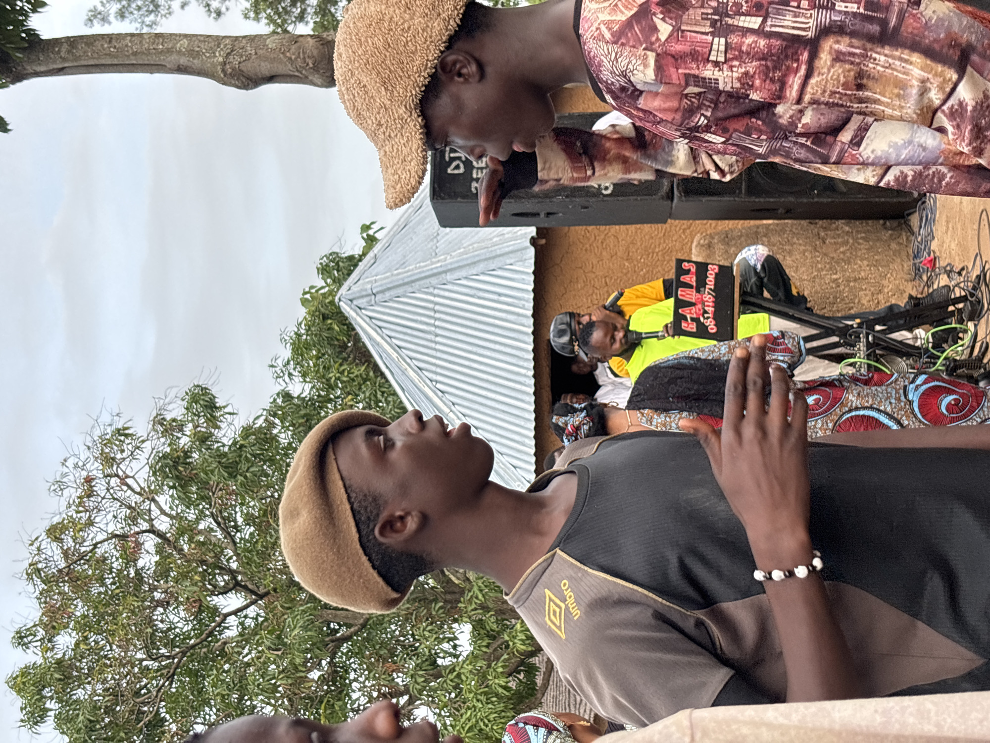 The image depicts an outdoor scene, possibly in Jos, Nigeria, where a group of young men are gathered. Two young men are in the foreground, facing each other in conversation. Both are wearing distinctive hats, one a brown beret and the other a textured, woven cap. The young man on the left is wearing a brown and black t-shirt with a yellow "umbro" logo. He is wearing a bracelet made of black and white beads. The young man on the right is wearing a patterned shirt with shades of red, pink, and brown.
In the background, a small, traditional-looking building with a corrugated metal roof is visible. A large black speaker is positioned next to the building. A DJ setup, including a laptop with "HAMAS" and a phone number displayed, is visible on a stand. Several other individuals are present in the background, some appearing to be part of a musical performance or event. The environment suggests a community gathering or outdoor event, with trees and a cloudy sky overhead. The ground appears to be dirt, with some electrical cables visible near the DJ equipment.