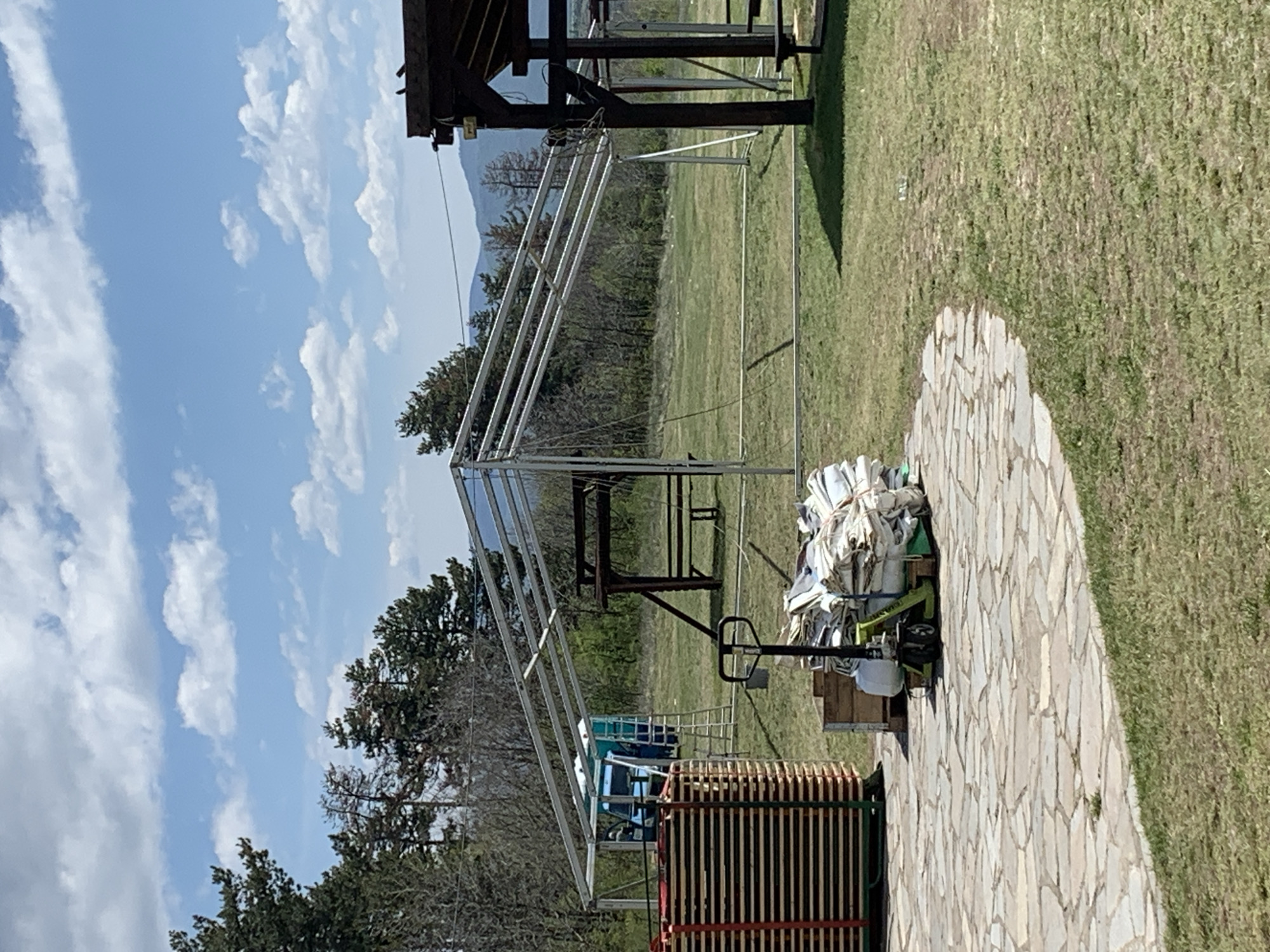 The image depicts an outdoor scene in Mirkovo, Bulgaria. In the foreground, a metal frame structure, possibly for a tent or awning, is partially erected. To the right, a pallet jack holds a stack of white bags and wooden crates on a stone-paved area that transitions to grassy ground. Further back, a wooden structure with a table and benches is visible behind the metal frame. In the background, a field with trees and a distant mountain range can be seen under a sky with scattered clouds. A blue vehicle is partially visible behind the wooden frame.