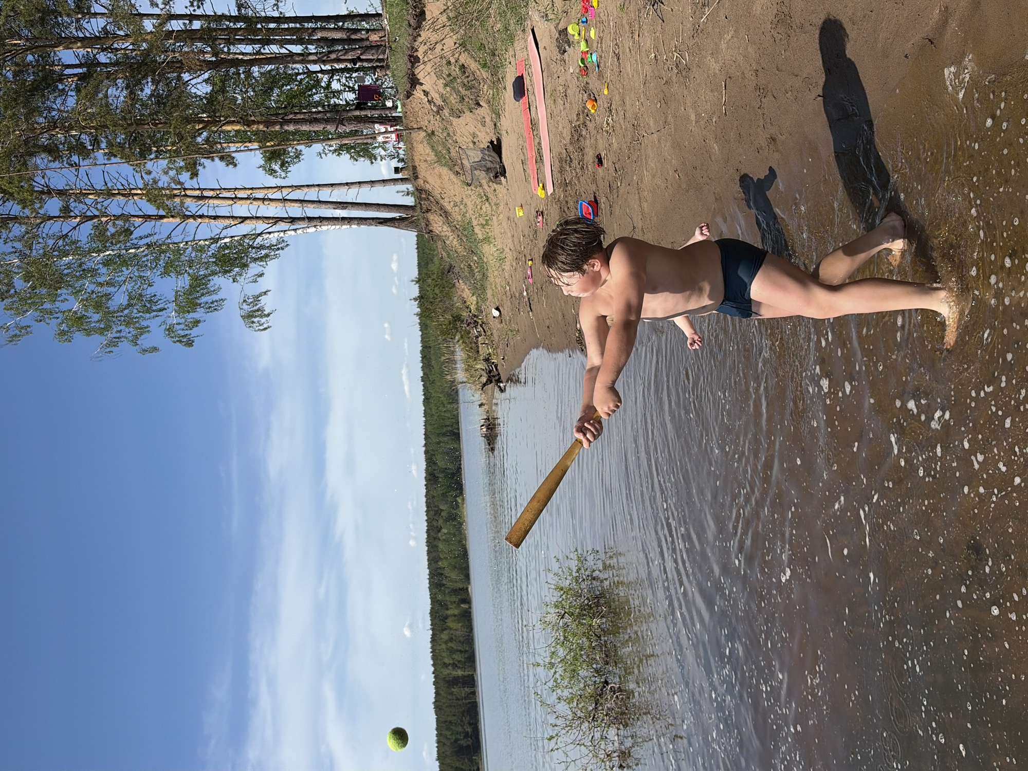 A boy is playing baseball at the edge of a body of water. He is standing in the water near the shore, holding a baseball bat in both hands, preparing to hit a tennis ball. The shoreline is sandy, with scattered toys and beach items. Trees line the shore beyond the sand. The sky is blue with some clouds. This is in Vekoshinka, Russia.