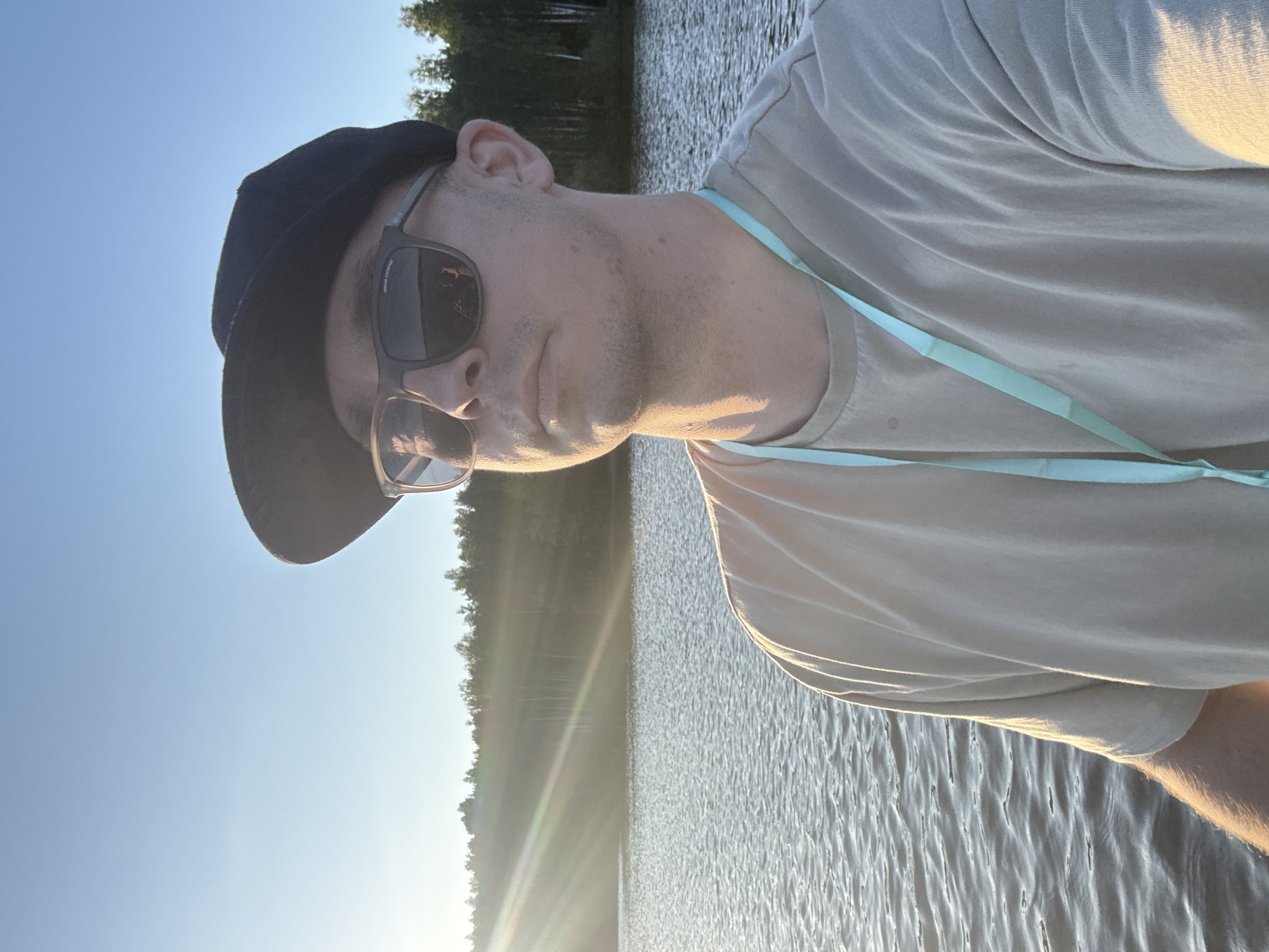 An individual wearing a black baseball cap and sunglasses is captured in a selfie outdoors. The person is standing on a body of water, likely a lake, with a forested shoreline in the background. Sunlight streams across the scene from the left, creating lens flare and illuminating the water. The person is wearing a light-colored t-shirt and a light blue lanyard is visible around their neck. The setting appears to be Gora (Khokhlovskoe S/P), Russia, given the sun's position and the natural landscape.