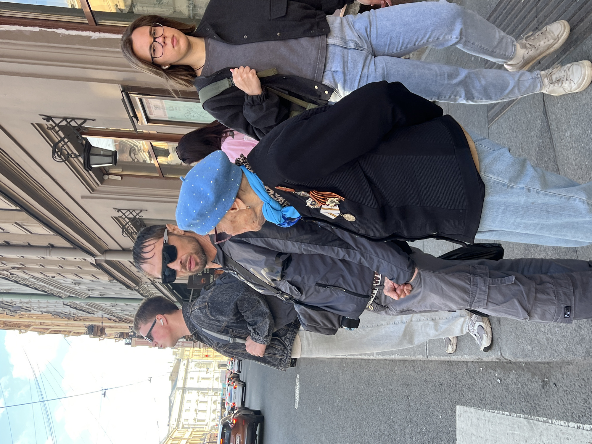 The image shows a group of people standing on a sidewalk in Saint Petersburg, Russia. From left to right, the first individual wears sunglasses, a gray denim jacket, white pants, and white shoes. The second person, also in sunglasses, is dressed in a gray jacket and gray cargo pants, and also wears New Balance sneakers. The central figure is an elderly woman wearing a light blue beret, medals, and a black jacket. Next to her is a person with long brown hair wearing a pink shirt. The last individual wears glasses, a dark jacket, a gray shirt, blue jeans, and white shoes. The backdrop includes buildings, part of the road, and cars.
