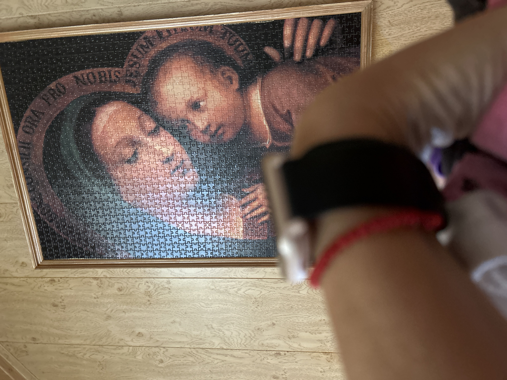 The image shows a framed puzzle of a religious artwork, featuring the Virgin Mary holding the infant Jesus. The puzzle is hanging on a wall with wood-paneled wallpaper. In the foreground, a person's arm and wrist are visible, wearing a black band and a red bracelet. The person appears to be holding a device, possibly a smartphone. The image does not contain any identifiable information about the city of Gelendzhik, Russia, or any specific location within it.