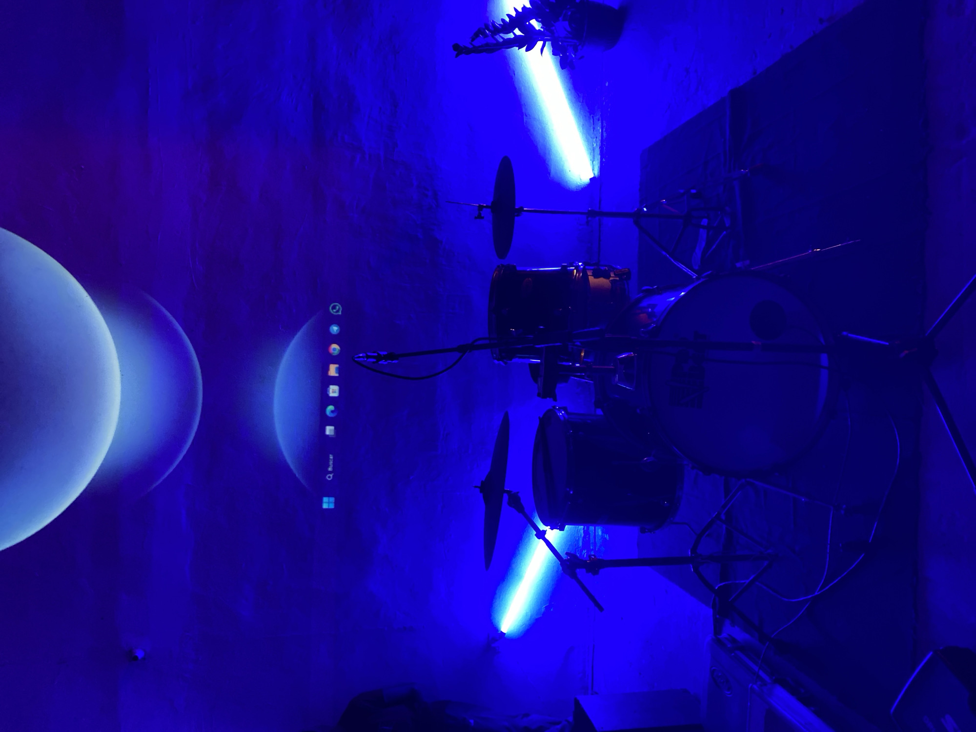 A dimly lit, blue-toned room houses a drum kit centered in the frame. The drum kit is positioned against a wall, with a light fixture above illuminating it. A computer monitor is visible on the left, displaying a screensaver with spherical shapes. The scene has a sterile and impersonal ambiance, with no visible individuals. The location is presumed to be in Tinaquillo, Venezuela.