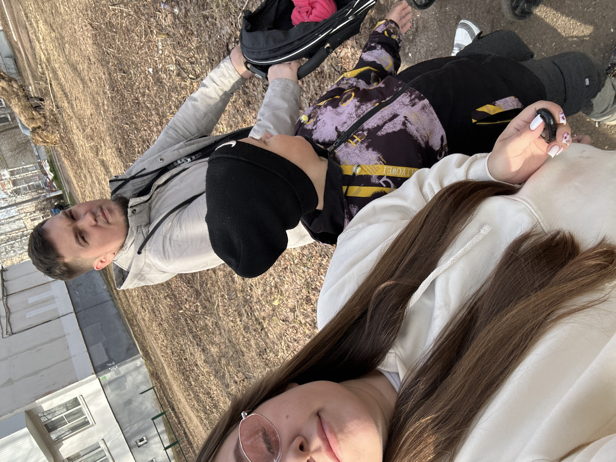 The image shows a selfie taken by a person with long brown hair, wearing rose-tinted glasses and a white hoodie. The person is lying down outdoors, with their head angled towards the camera. Behind them is another person, a male, wearing a light grey jacket and a black beanie. He is looking at the camera. There is a child in a black and purple jacket lying between them. A black bag is also visible, along with some other people's legs and feet in the background. The ground appears to be covered with dry grass or leaves. In the background, there is a building with a fence. Based on this detail and the presence of deciduous trees, the location appears to be Chaikovsky, Russia.