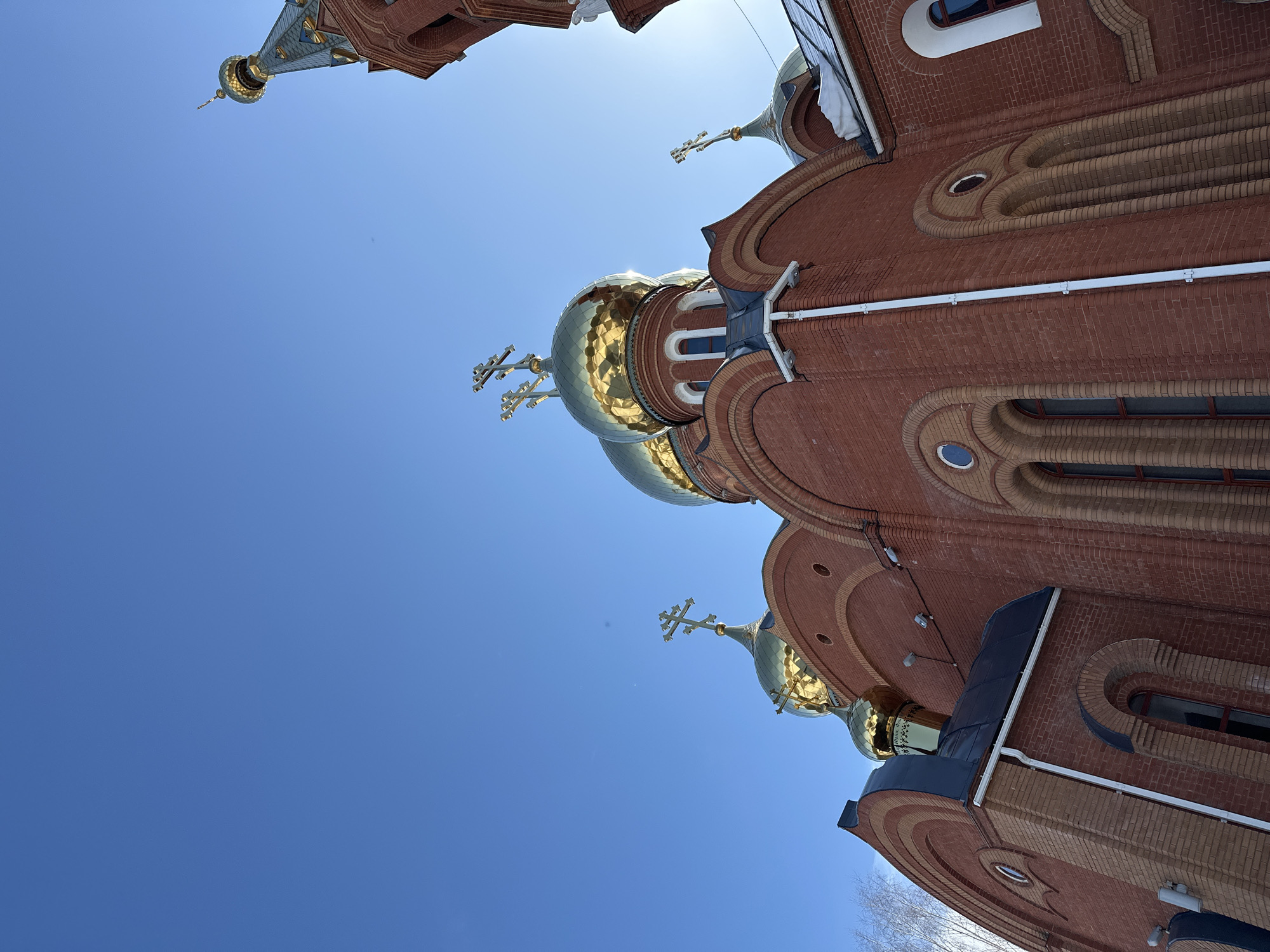 The image displays the Nikolsky Cathedral (Cathedral of St. Nicholas) in Chaikovsky, Russia, under a clear, deep blue sky. The church is constructed from red brick, with lighter reddish-brown bricks used for decorative banding and arched details around windows. Five golden onion domes are visible, each crowned with a golden triple-bar Orthodox cross. Two large golden domes are prominent in the central frame, with a smaller golden dome visible to their left. A bell tower featuring a patterned green and blue conical spire with a cross is partially visible on the far right. Windows are primarily tall, narrow, and arched, although a small circular window (oculus) is also present. Dark, sloped roof sections are visible beneath the domes, and white gutters and downspouts are affixed to the brick walls. Patches of snow are evident on some roof ledges. Direct sunlight illuminates the church from the right, causing reflections on the golden domes and a slight lens flare. Bare tree branches are visible on the extreme left. No individuals are depicted. The photograph is captured from a low-angle perspective, looking upwards at the cathedral.