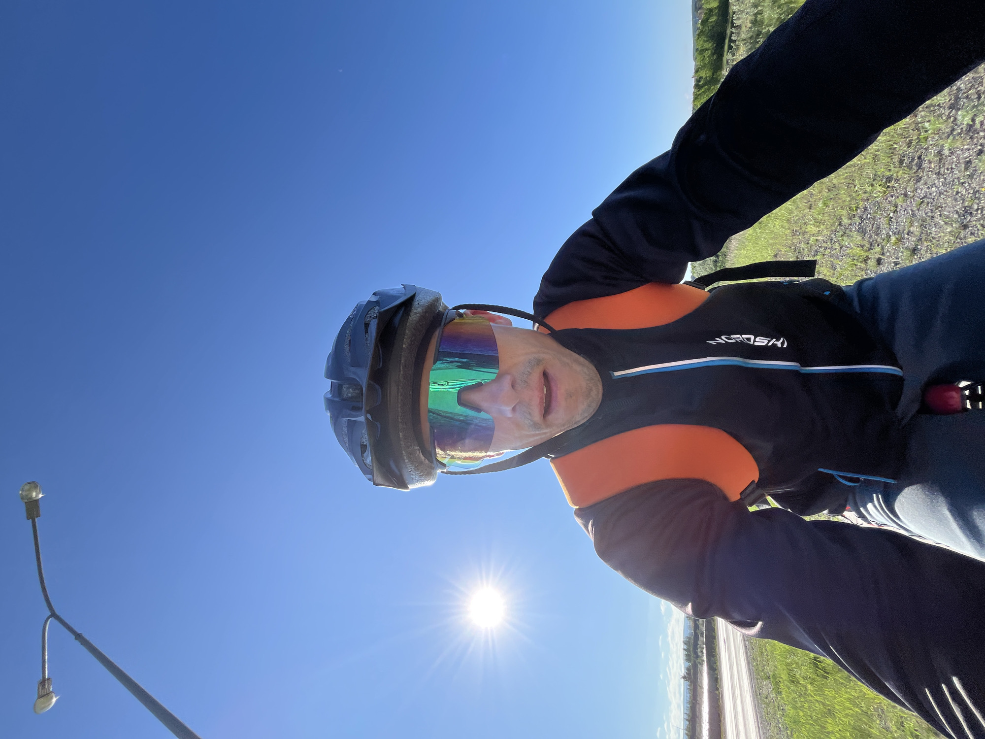 A first-person perspective image shows a person wearing a bicycle helmet and reflective sunglasses. The person is also wearing a black cycling jacket with an orange vest over it, and dark cycling shorts. The background shows a clear blue sky with the sun shining brightly, indicating a sunny day. To the left, a street lamp with two lights is visible. The lower part of the image shows a grassy roadside next to a paved road, with some trees in the distance. The image appears to have been taken while the person is cycling, as suggested by the low angle and the blurred motion of the surroundings. The specific location in Chaikovsky, Russia, cannot be determined from the visual information provided in the image alone.
