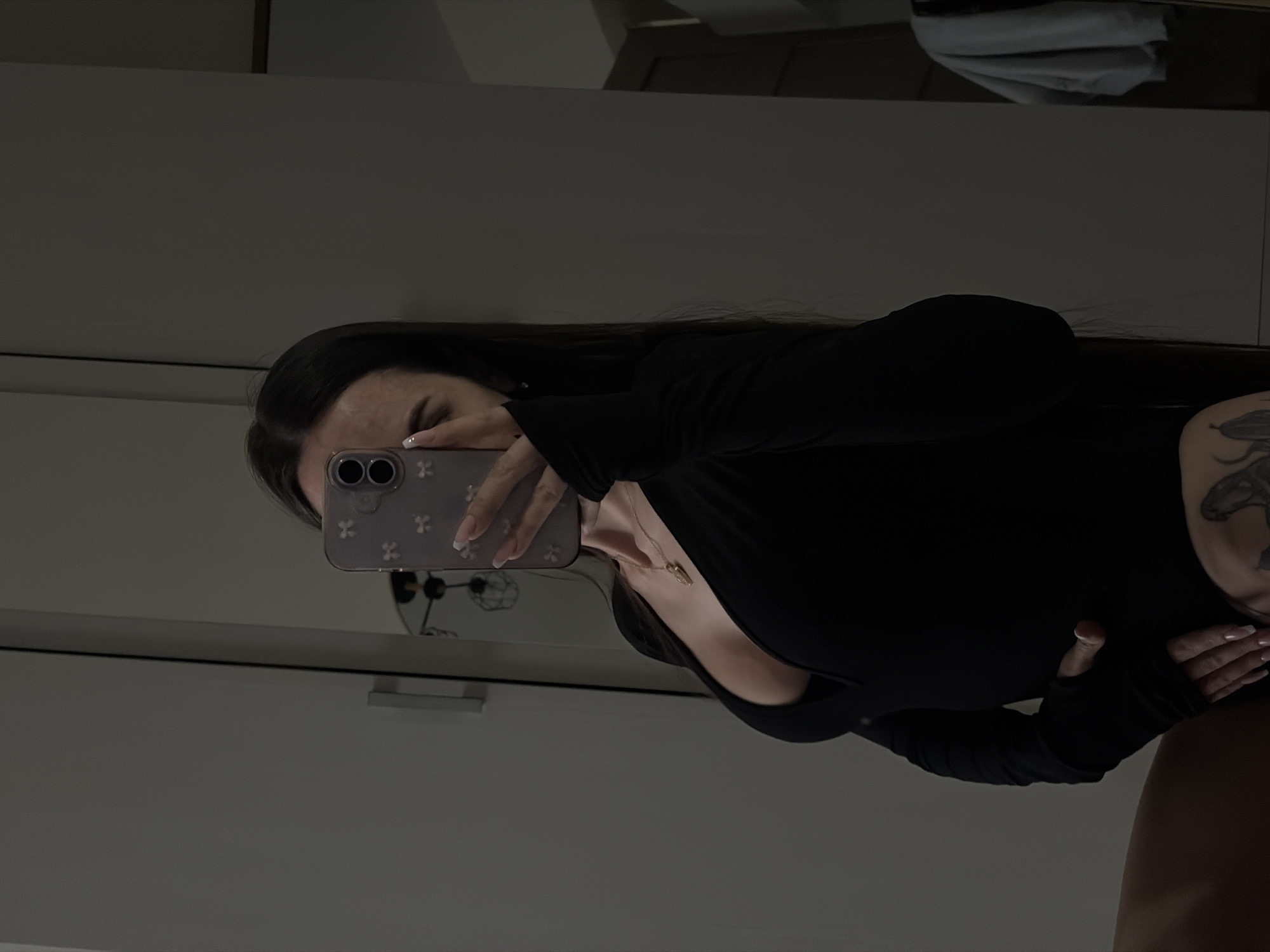 A person is captured taking a mirror selfie in an indoor setting. The subject, likely female, has long dark hair and is wearing a black, fitted, long-sleeved top or bodysuit with thumbholes. A delicate necklace with a small, light-colored pendant hangs around her neck. A tattoo featuring dark, intricate designs is visible on her upper right arm/shoulder. Her fingernails are manicured with light-colored polish, possibly a French manicure style.
The person holds a light-colored smartphone with a decorative case adorned with small, cross-like patterns. The phone covers most of her face, with only a portion of her forehead and eyes visible above it. The phone's two rear camera lenses are clearly seen. The reflection in the mirror reveals a room with a light-toned wall or cabinet behind the subject. Faint reflections of what appears to be folded fabric or towels can be seen in the upper background of the mirror. The lighting is subdued, creating a soft, indirect atmosphere. The image is oriented sideways, with the subject positioned diagonally across the frame. No discernible text is visible within the image. The implied location is Saint Petersburg, Russia.