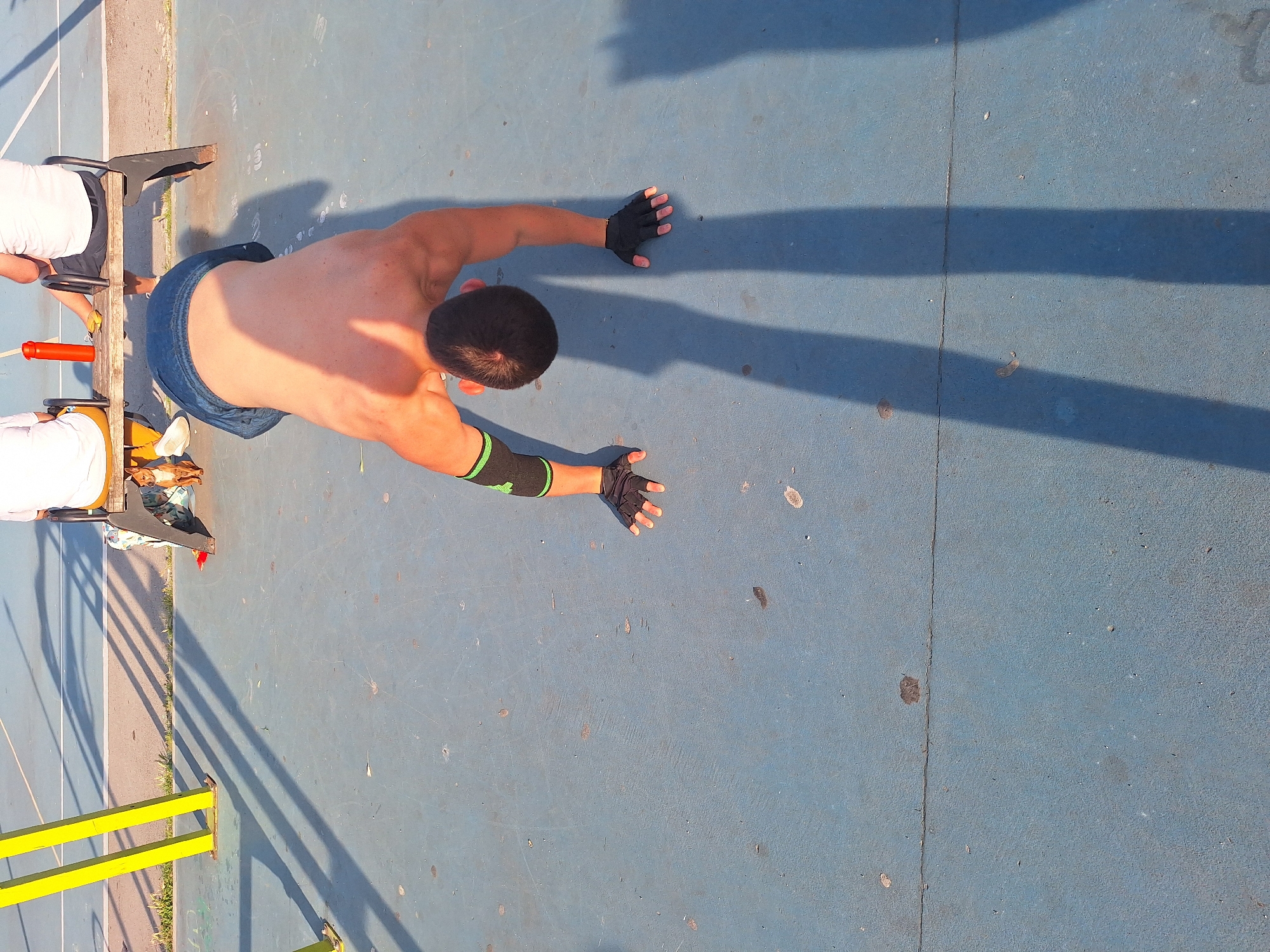 An aerial shot captures a man in a push-up position on a blue outdoor court, with his body casting a long shadow. He is shirtless, wearing blue denim shorts and black fingerless gloves with green accents on his forearms. His hands are extended forward on the ground, palms down, fingers spread. His head is turned slightly to the right, revealing dark hair.
To the left of the man, a wooden structure with metal supports is visible. Two individuals, partially visible, are seated on this structure, wearing white tops. An orange object, possibly a piece of equipment, is also on the structure. Yellow bars of a fence or railing are in the foreground. The court appears to be a sports facility, possibly a basketball or tennis court, with faint lines visible on the surface. Grass can be seen growing along the edge of the court.
The lighting suggests it is a sunny day, likely late afternoon or early evening given the long shadows. The overall mood is one of physical activity and training, possibly in a public park or sports complex in Varna, Bulgaria. There is no visible text in the image.