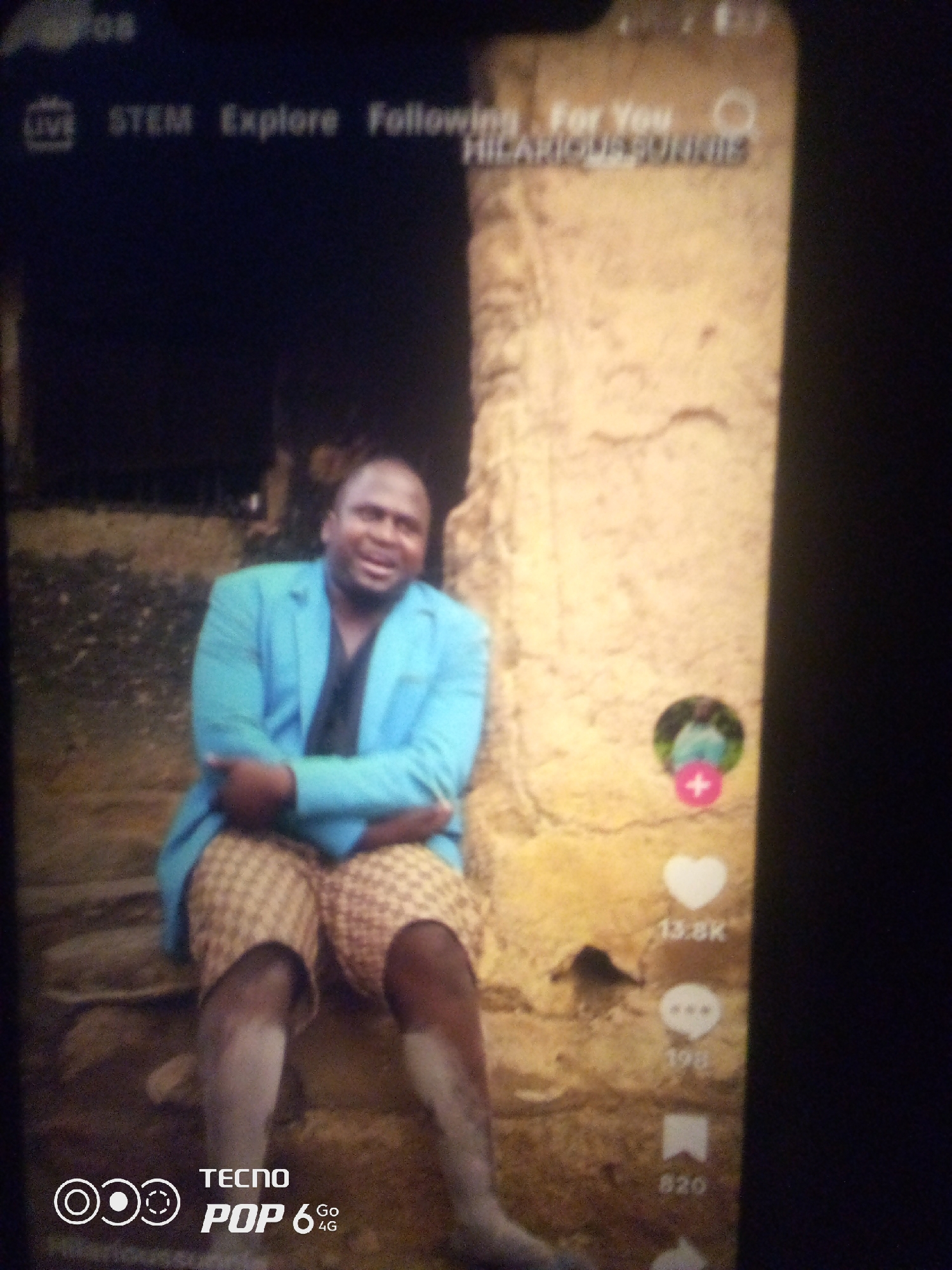 In a digitally captured image, a man wearing a light blue suit jacket and checkered shorts is seated against a textured, weathered wall. His expression is jovial as he looks slightly off-camera. To the right of the man, a series of icons are visible, including a heart with a count of "13.8K", a speech bubble icon, and a bookmark icon with a count of "820". The background appears to be outdoors, with a dark, indistinct area behind the man that might suggest nighttime or a shadowed enclosure. The image displays digital interface elements at the top and bottom, including text that reads "LIVE", "STEM", "Explore", "Following", "For You", "HILARIOUS FUNNIES", and a "TECNO POP 6 4G" logo. The environment does not provide specific landmarks to pinpoint a location within the city of Howayi, Nigeria.