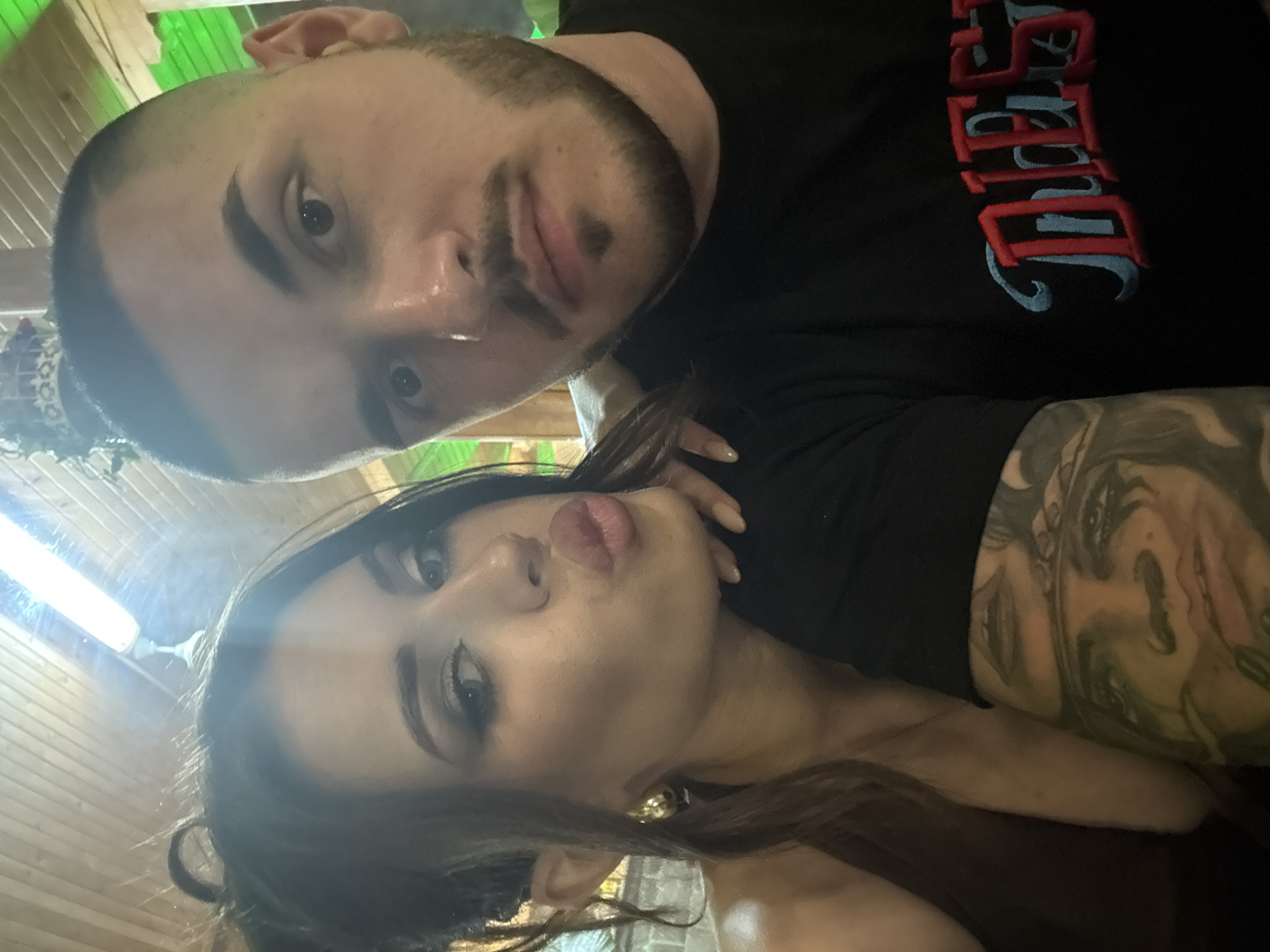 A close-up, waist-up selfie depicts a man and a woman in an indoor setting, identified as Debrashtitsa, Bulgaria. The woman, positioned on the left and slightly behind the man, has long dark hair styled in a partial updo with strands framing her face. She wears noticeable eye makeup, including winged eyeliner and defined eyebrows, and her lips are pursed in a "kissing" gesture with dark lipstick or gloss. A large, gold-toned hoop or disc earring is visible on her left ear. Her attire appears to be a dark brown or bronze halter-neck garment. Her right hand, with light-colored manicured nails, rests on the man's left shoulder.
The man, centrally positioned to the right of the woman and closer to the camera, has a close-cropped dark fade haircut and a neatly trimmed dark beard with a distinct 'soul patch' below his lower lip. He wears a black t-shirt with a partially visible embroidered logo on the chest in red and light blue, appearing to spell "DIESEL". An extensive, detailed sleeve tattoo, depicting stylized faces and figures, covers his left arm (visible on the right side of the image). He looks directly at the camera with a neutral expression.
The background features a wooden plank ceiling with a bright white fluorescent tube light fixture visible towards the top. Green accent lighting illuminates the upper right side of the frame, indicating artificial lighting. A textured wall, possibly brick or stone, is visible behind the woman. A decorative object, resembling a small, ornate crown or plant fixture, hangs from the ceiling near the top center. The overall scene suggests a casual social interaction, possibly in a restaurant or bar environment.