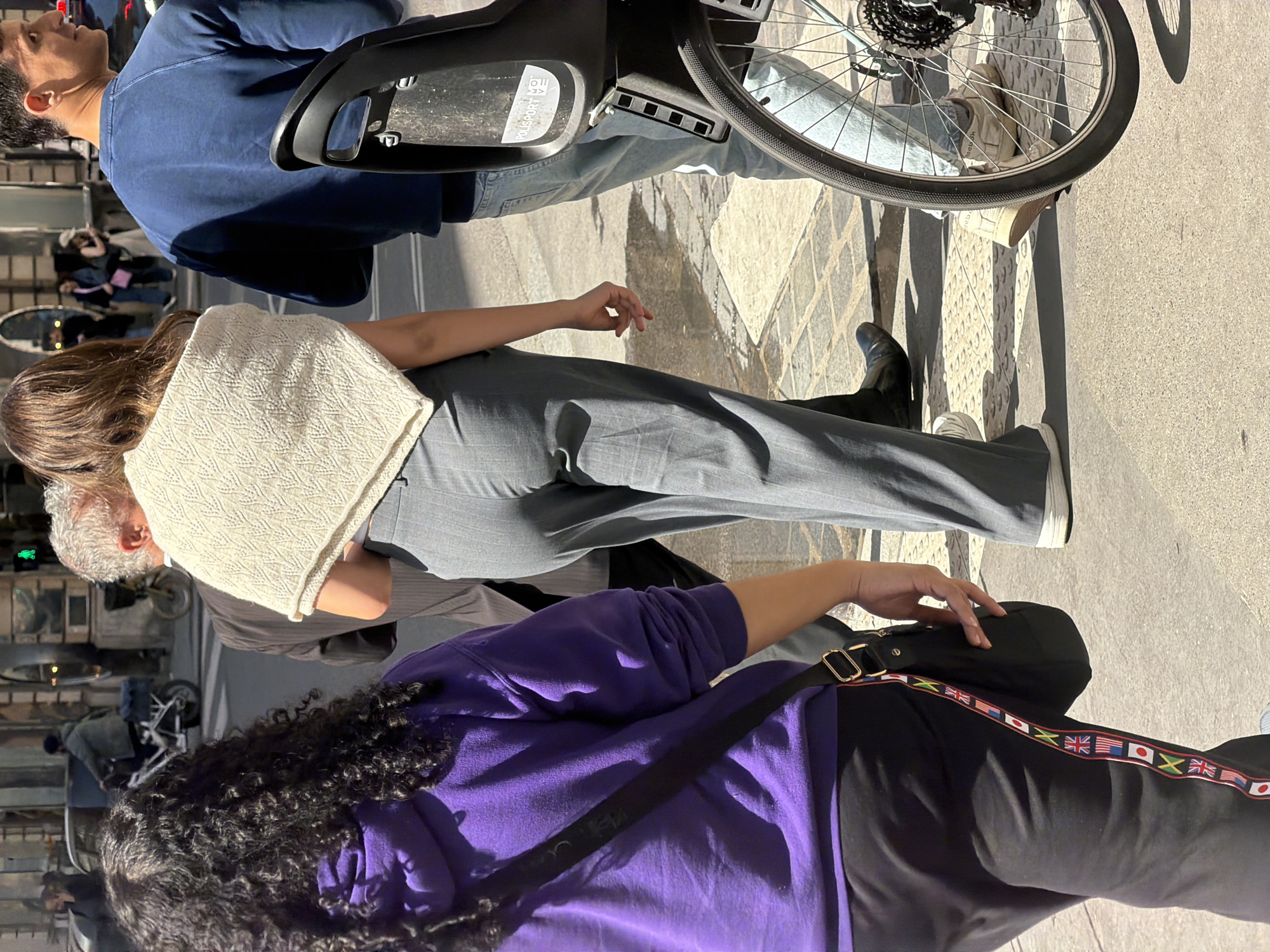 The image shows a street scene. A man in a blue shirt is standing next to a bicycle. Nearby, a woman in a grey outfit and a beige sweater is visible. In the foreground, another person with curly hair wearing a purple shirt is also present. The pants of the person in purple have a strip decorated with international flags. The ground consists of interlocking pavers. The scene is likely in a city, but it is not clear from the image alone whether it is in Paris.