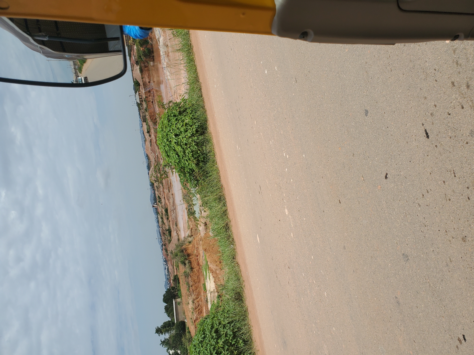 The scene depicts a landscape in Reifield-Ist, Nigeria, observed from the interior of a vehicle, likely a tricycle, positioned on a paved road. The perspective is angled slightly upwards and to the left.
In the foreground, a light grey-brown asphalt road occupies the bottom half of the image. On the far right, a vertical yellow structural element of the vehicle and a lower grey interior panel are visible. A black-framed rearview mirror is prominently positioned in the upper right, reflecting a road behind the vehicle and a distant white vehicle (possibly a bus or van) approaching.
The midground features green bushes and grass lining the left edge of the road, beyond which the terrain descends into an extensive area of reddish-brown, disturbed earth, indicative of excavation or significant erosion. Within this area, several irregularly shaped, reddish-brown water bodies are visible. Further in the mid-distance on the left, a few small, simple buildings, one with a light-colored roof, are situated on higher ground.
The background consists of a uniformly overcast, light grey sky. The distant horizon reveals faint outlines of rolling hills or elevated land, sparsely dotted with structures, suggesting a peri-urban or transitioning rural environment. No individuals are directly visible in the landscape. The overall environment suggests an area undergoing land development or impacted by natural processes in a tropical setting.