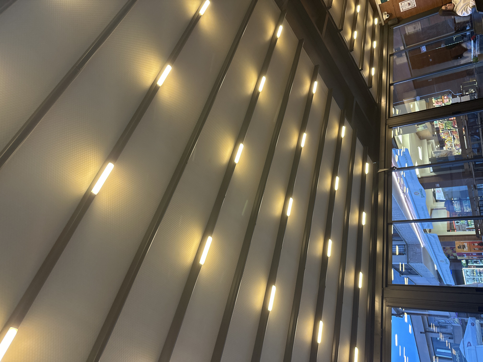 The image depicts an enclosed outdoor seating area, likely part of a cafe or restaurant, at dusk or early evening. The overhead structure is a light-colored, textured retractable awning or roof system, supported by dark metal beams. Numerous rectangular, warm-toned LED light fixtures are evenly spaced across the ceiling, illuminating the space. The area is enclosed by large glass panels on the lower perimeter.
Through the glass, an urban street scene in Plovdiv, Bulgaria, is visible. To the left, a modern building with multiple windows and a partial white canopy marked "Copart" is seen under a bright blue sky. To the right, another building features a storefront with a prominent blue awning displaying text, including "КАМЕНИЦА" (Kamenitza) and "БИРА" (Beer), indicating a Bulgarian location. Below this awning, the brightly lit interior of a store, possibly a convenience market, is visible with various products on shelves.
Inside the seating area, on the far right, a woman with dark hair wearing a light-colored top is seated, partially visible from the chest up, looking towards her right. A wooden-paneled wall section is behind her. A white CCTV security camera is mounted in the upper right corner of the indoor ceiling. Dark furniture, consistent with tables and chairs, is reflected in the glass panels. No specific actions or interactions are discernible beyond the woman's passive presence.