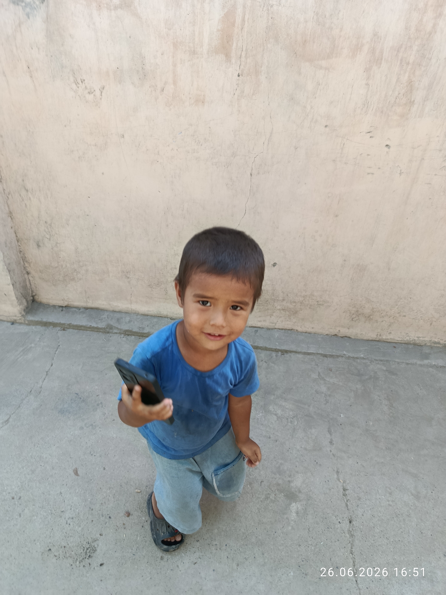 A young boy, likely a child, stands in front of a textured, light-colored wall. He is wearing a blue t-shirt and light-colored shorts. In his right hand, he holds a black smartphone. The boy is looking directly at the camera with a slight smile. His attire and the background suggest an outdoor setting, possibly a courtyard or alleyway with a concrete floor and a weathered wall. The lighting indicates it is daytime. A date and time stamp in the bottom right corner reads "26.06.2026 16:51".