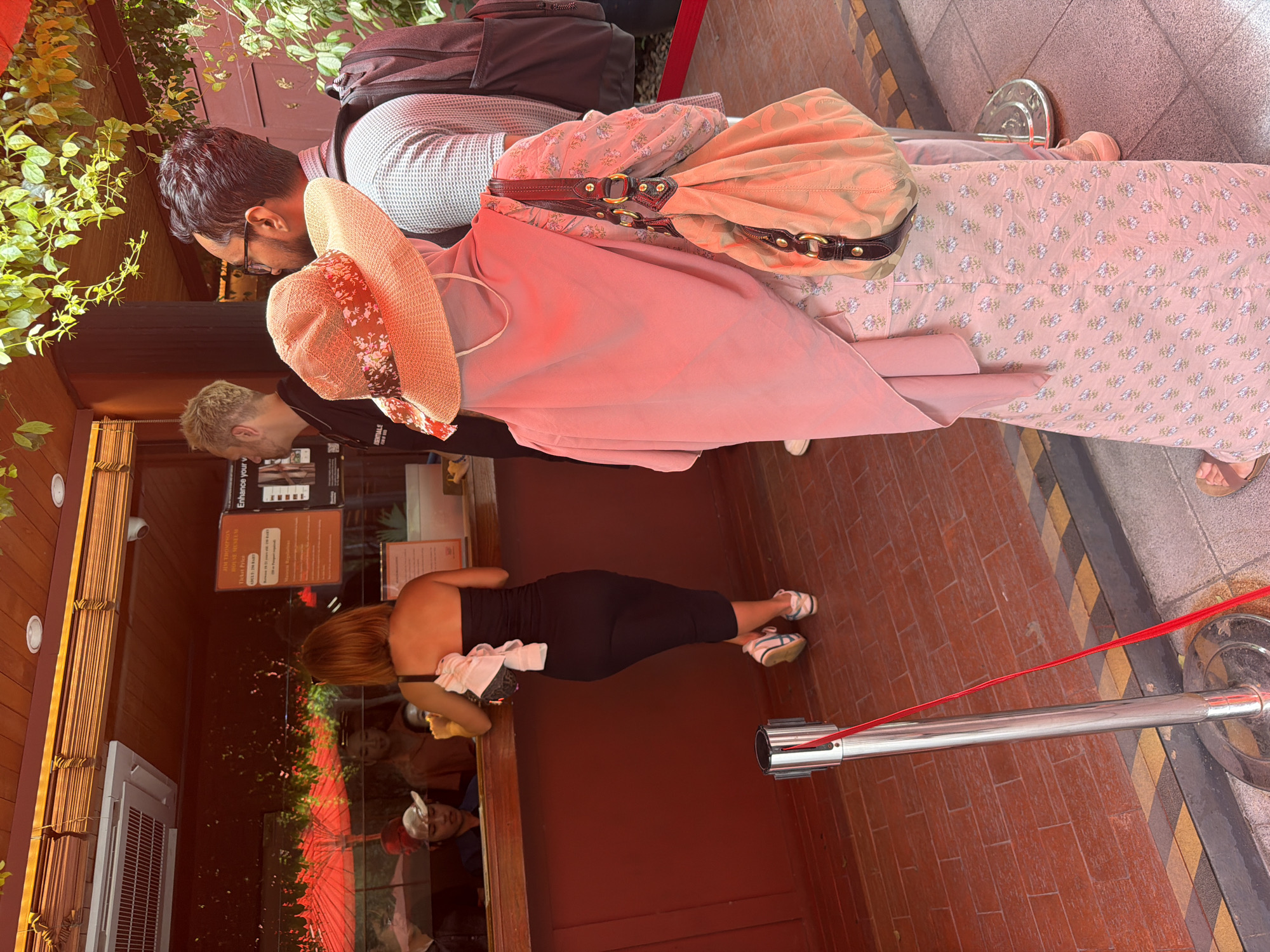 The image shows a group of people at a ticket counter, possibly at a tourist attraction in Bangkok, Thailand. A woman in a black strapless dress is at the counter, seemingly interacting with an employee. Behind her, a person wearing a pink hijab and a floral dress, and another person with glasses and a backpack are waiting in line. The background reveals a red wooden structure with windows, a sign displaying "Jim Thompson Museum Ticket Price", and reflections of red umbrellas and people. The environment appears to be outdoors, with greenery visible above. A stanchion with a red ribbon is positioned in the foreground, indicating a queue.