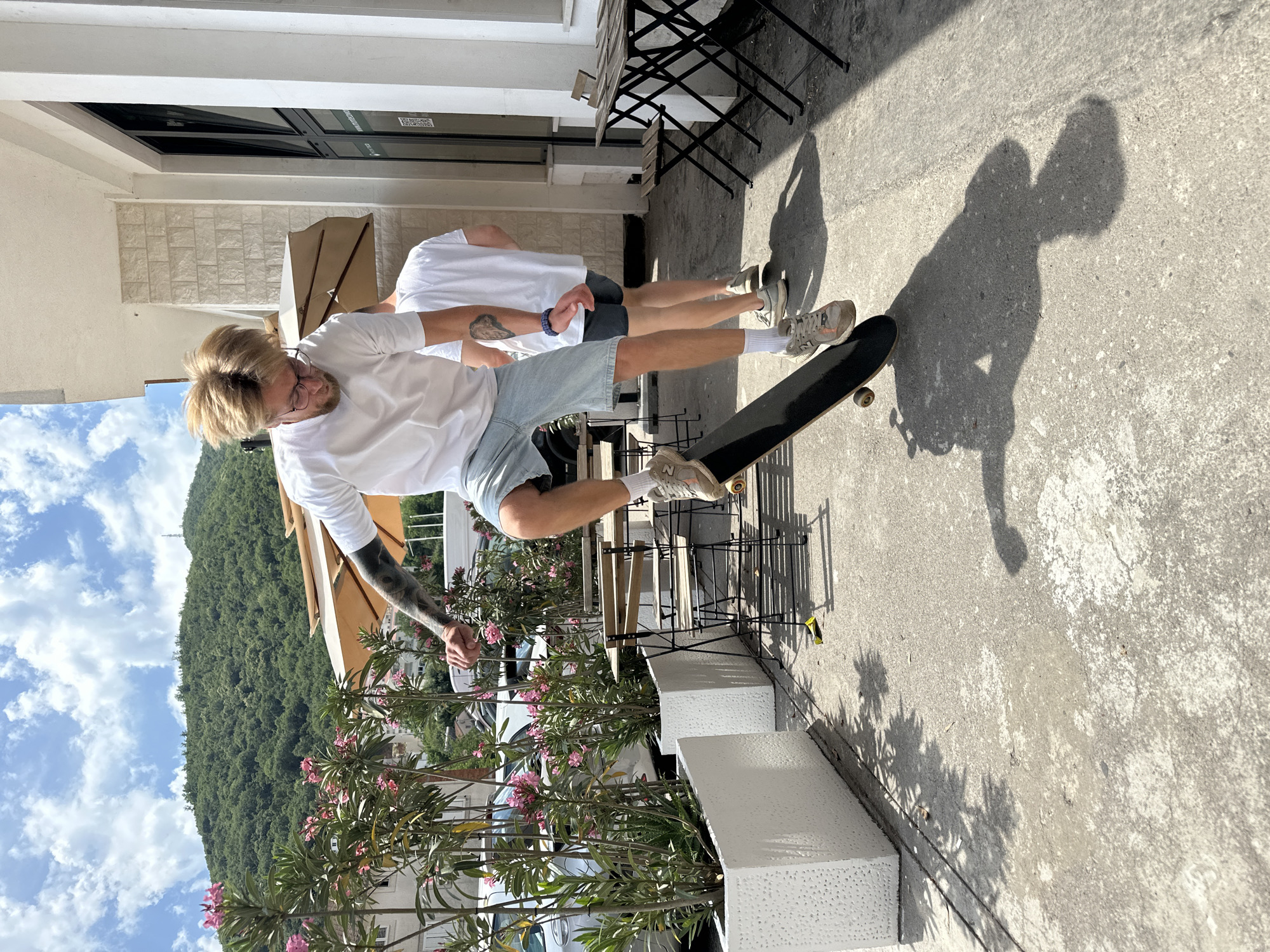 In Budva, Montenegro, on a sunny day, a young man with blond hair and glasses is skateboarding. He is wearing a white t-shirt and light blue denim shorts, with his skateboard mid-air as he performs a trick. Another person in a white t-shirt stands behind him. The scene takes place outdoors on a concrete sidewalk, next to a building with white stone cladding and large windows. In the background, there are potted plants with pink flowers, parked cars, and a lush green hill under a blue sky with scattered clouds. Outdoor cafe tables and chairs are also visible.