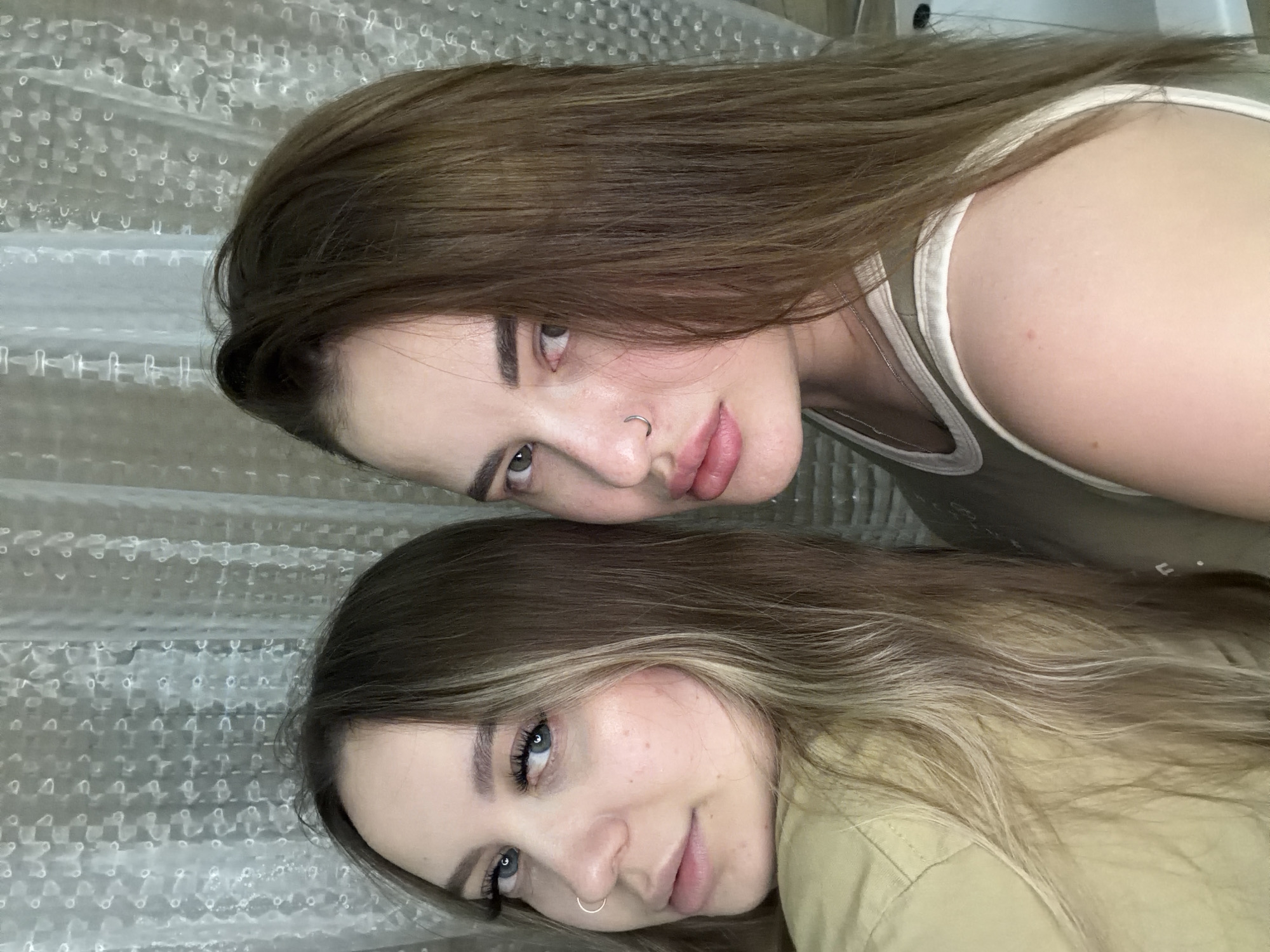 Two women with brown hair are lying on their sides on top of clear bubble wrap. The woman on top has medium brown hair, dark eyebrows, a nose ring, and is wearing a tank top. The woman on the bottom has lighter brown hair with blonde highlights, blue eyes, a nose ring, and is wearing a collared shirt. The location appears to be indoors with the bubble wrap as a backdrop. There is no indication of Krasnodar, Russia.