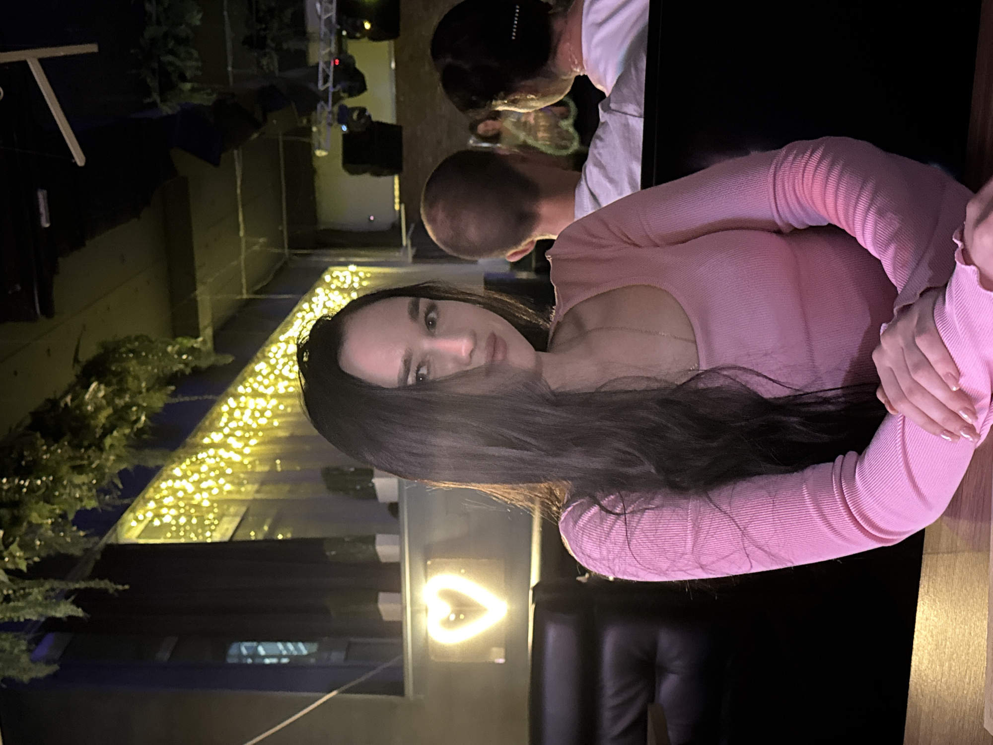 The image shows a woman with long, dark hair wearing a pink, ribbed, long-sleeved shirt. She is seated in what appears to be a dimly lit establishment, possibly a bar or restaurant. In the background, there are other patrons, with one man's bald head visible and another person's back and head turned away. The environment features hanging greenery, string lights, and a neon heart-shaped light. The scene is set at night, with artificial lighting creating a warm ambiance. The location is identified as Novosibirsk, Russia.