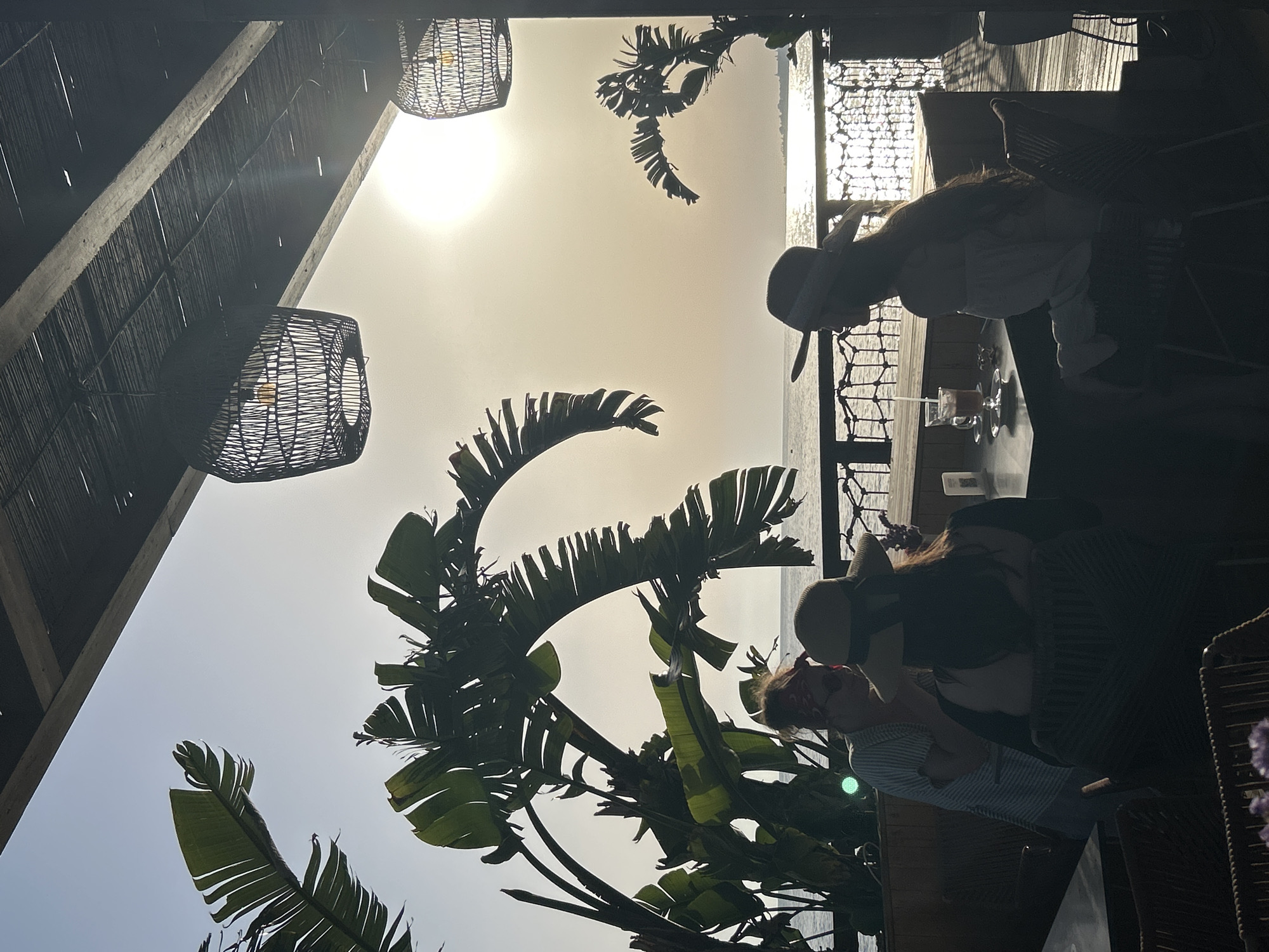 The image depicts an outdoor cafe setting in Alanya, Turkey. Two people are seated at a table, one wearing a hat. They appear to be on vacation or leisure time. A drink with a straw is visible on the table. Lush green tropical plants frame the scene, with the sun shining through the leaves. Woven lamps hang from the roof of the outdoor seating area. The sky is bright, and the sea is in the background.