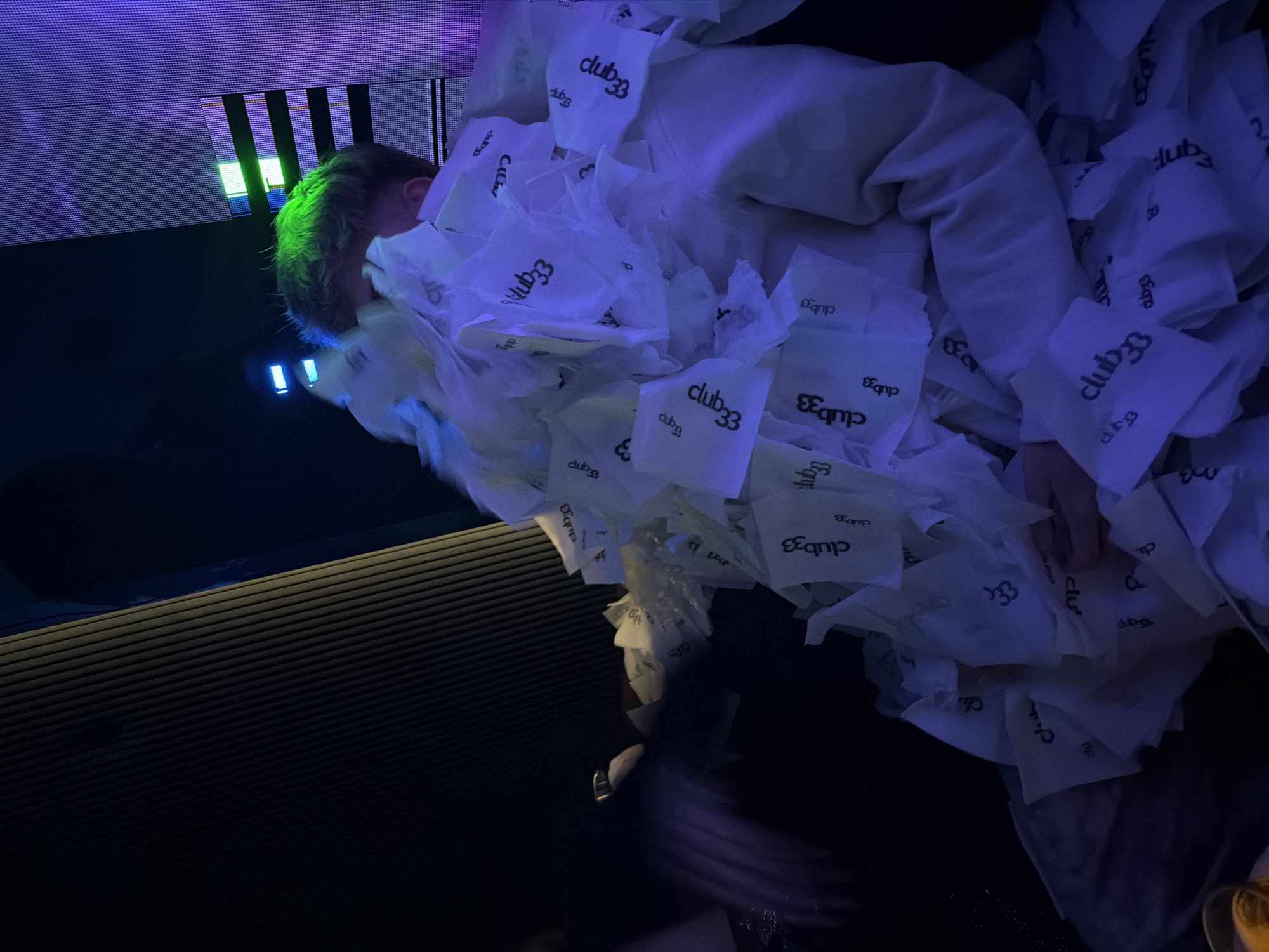 A person with green hair is lying on a surface, almost entirely covered in white napkins with the inscription "club33" on them. The person is wearing a white sweater. The surrounding environment appears to be a dark interior, illuminated by blue and green lights. A ribbed surface is visible nearby, and an electronic screen is in the background. The city of Sofia, Bulgaria is not indicated in the image.