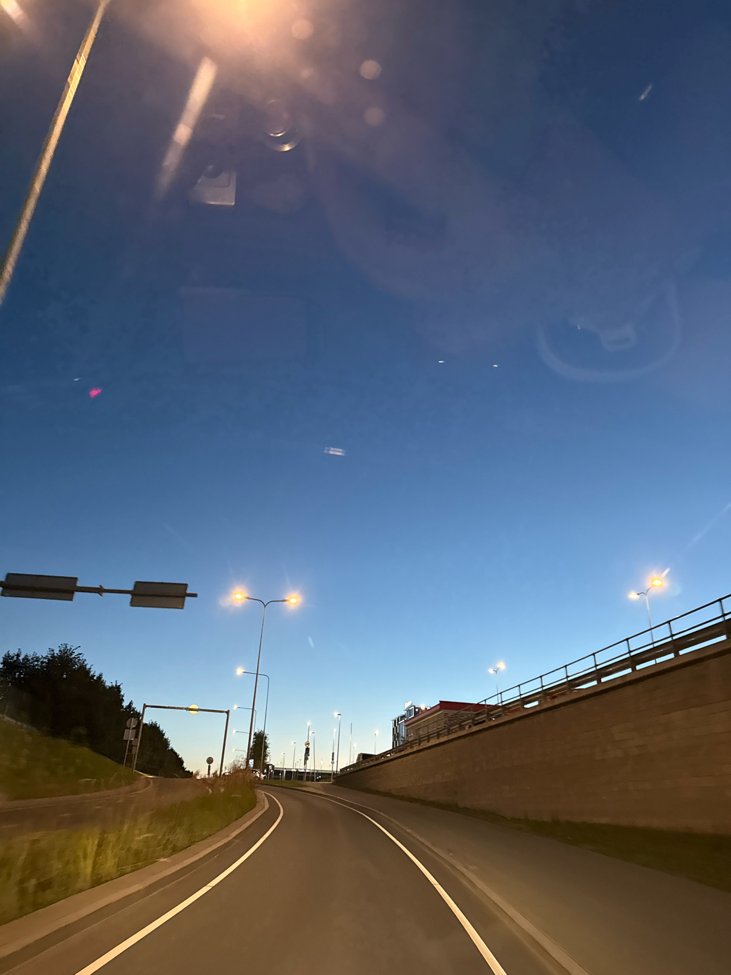 The image depicts a perspective from a vehicle traveling on a road in Tallinn, Estonia, during twilight. The sky is a clear, deep blue, transitioning from the fading light of day. Streetlights illuminate the road and surrounding areas.
On the left side of the road, a grassy embankment rises, with trees and vegetation visible in the distance. A road sign with two rectangular panels is positioned above the road. Further down the road, a traffic light can be seen.
To the right, a raised structure, possibly an overpass or bridge, is lined with a metal railing. The wall of this structure is made of concrete or stone blocks. A red-roofed building or a truck with a red structure is visible atop the overpass. Multiple streetlights are mounted along the overpass, their bright lights contrasting with the darkening sky.
The road itself is paved and marked with white lines. The perspective suggests the vehicle is navigating a curve. Reflections are visible on what appears to be the windshield of the vehicle, including a bright light source at the top of the frame and circular patterns. The overall scene captures a common urban driving environment as night begins to fall in Tallinn.