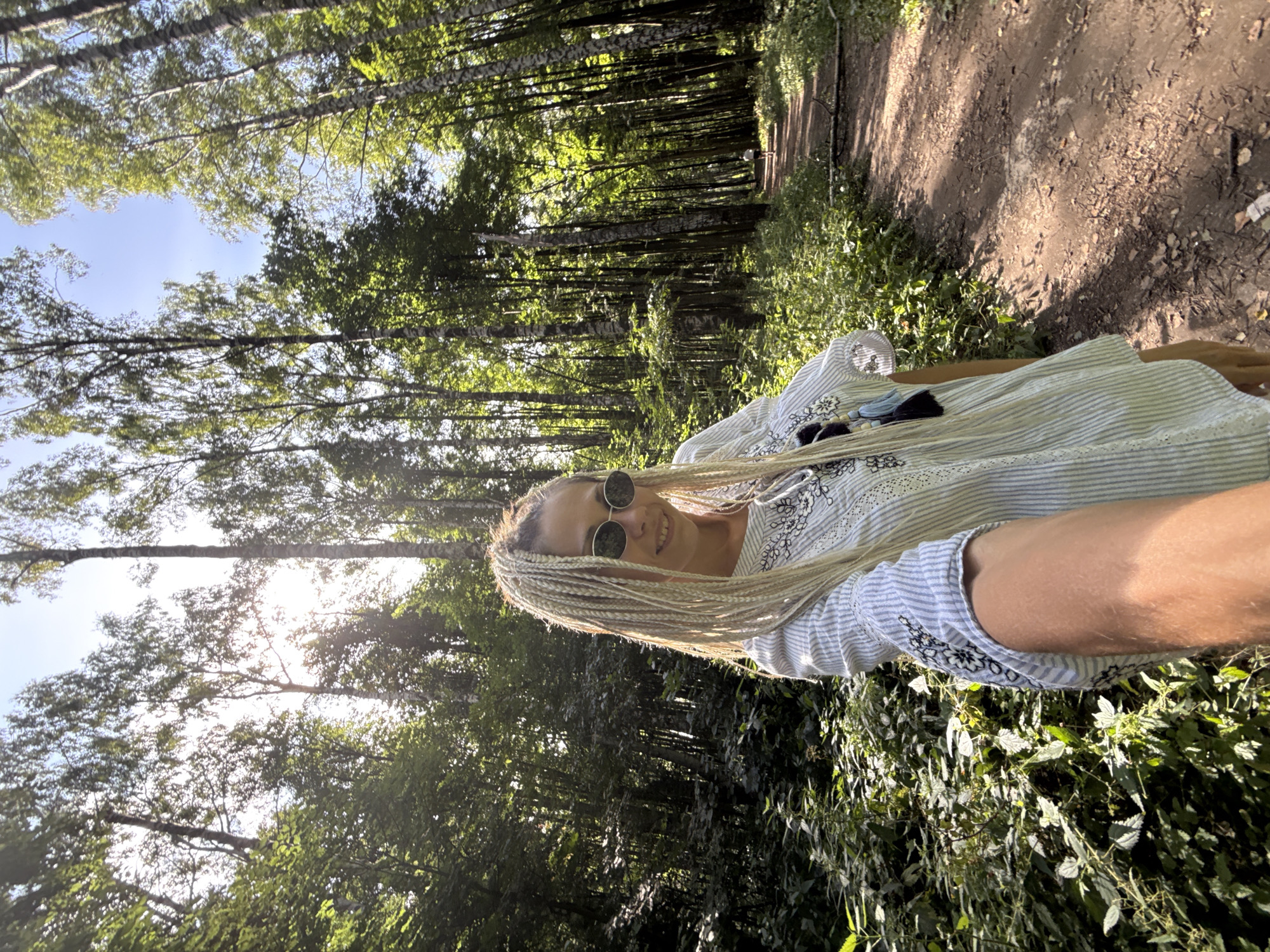 A woman with blonde braids and sunglasses poses for a selfie in a forest on a sunny day. She is wearing a white striped shirt with black embroidery. The forest path is visible in the background, with trees lining both sides. The image appears to be taken in Dumino, Russia.