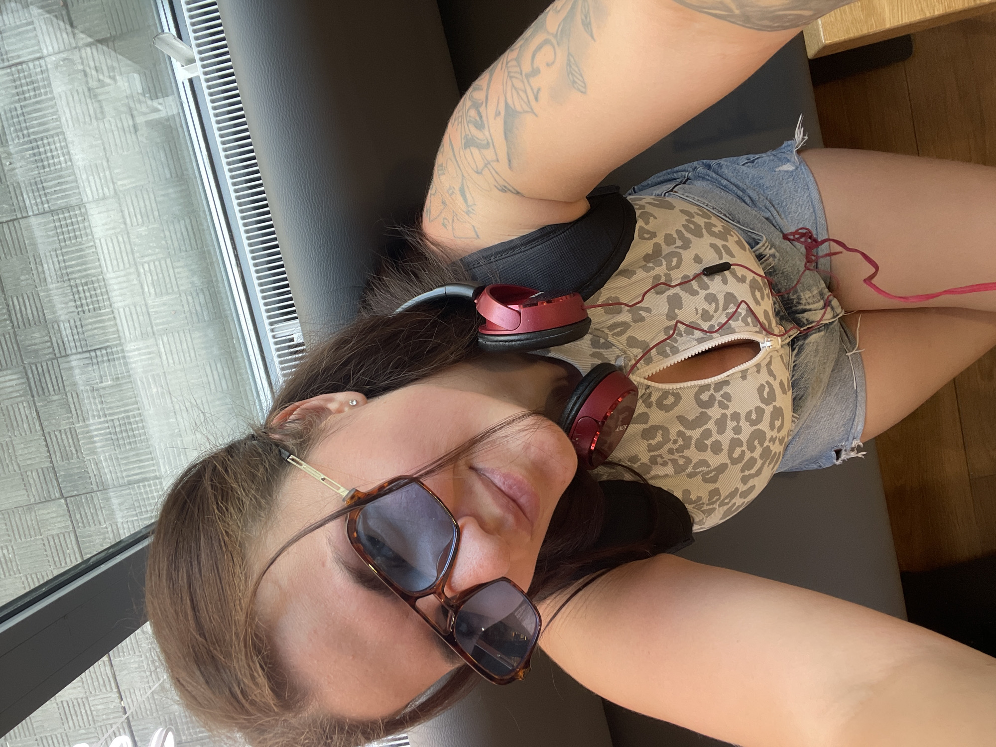 A woman with brown hair is lying down on a gray couch, wearing sunglasses and headphones. She is wearing a tan animal print tank top and denim shorts. Her left arm is visible, featuring a tattoo. A window is visible to the left. The location in the city of Misaylovo, Russia cannot be confirmed.