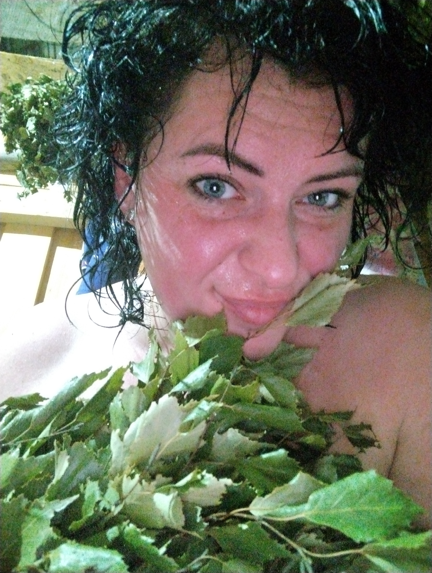 This close-up image features an adult woman, likely in a sauna or banya setting in Grishanki, Russia. Her face is flushed and appears moist from heat or steam, with droplets visible on her skin and wet, dark, curly hair. She has blue eyes and a slight, content smile. A large bundle of fresh, green leafy branches, resembling a venik (a traditional sauna besom), is held up to her face and chest, partially obscuring her mouth and body. In the background, wooden planks of what appears to be sauna benches are visible. The overall atmosphere suggests a moment of relaxation or enjoyment during a banya session. No text is visible in the image.