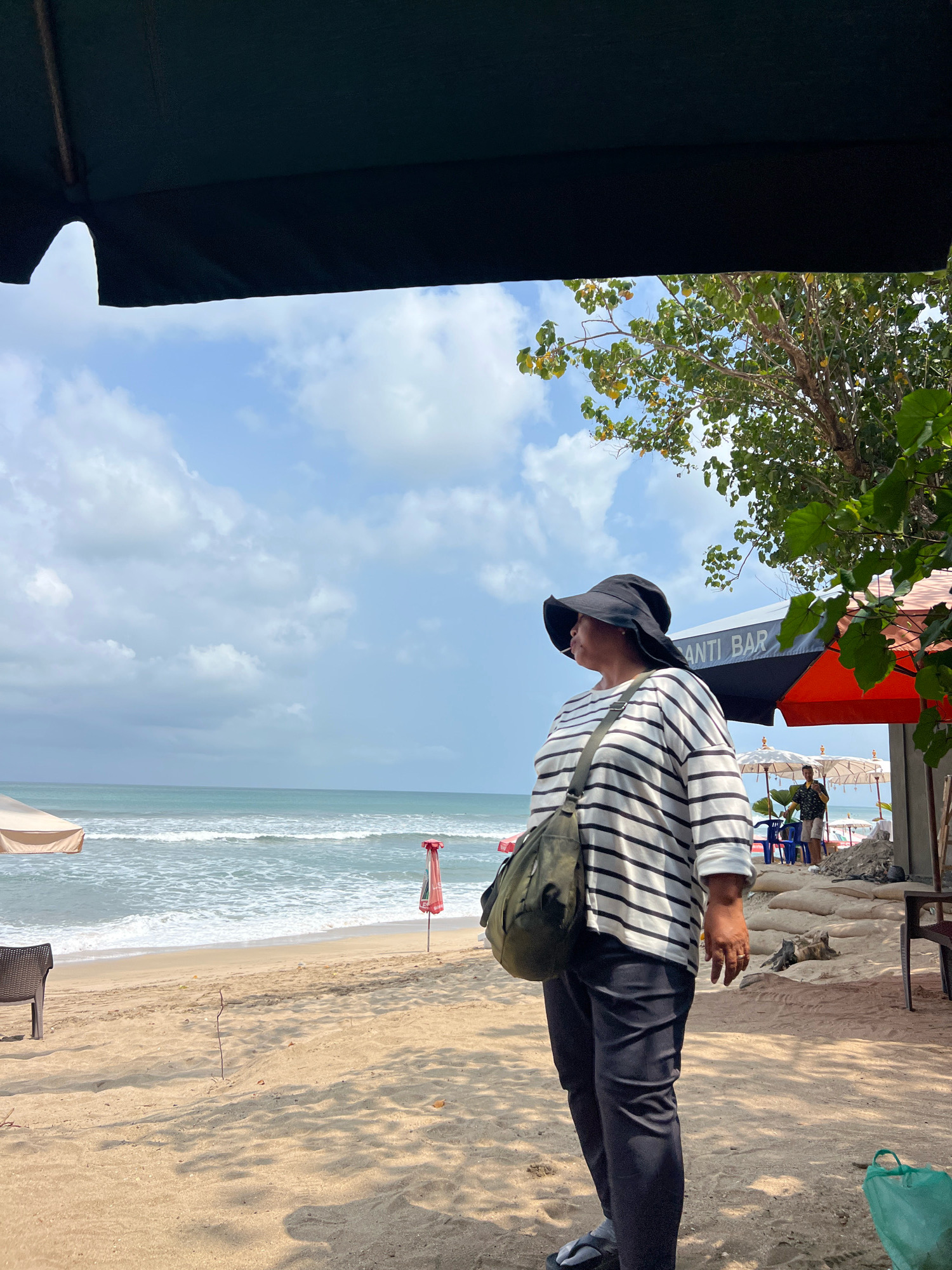 A woman wearing a black wide-brimmed hat and a black and white striped long-sleeved shirt stands on a sandy beach in Kuta, Indonesia. She has a green bag slung across her body and is wearing dark pants. The woman is looking out towards the ocean, which has gentle waves. In the background, there are beach umbrellas, a bar labeled "ANTI BAR," and a few people. Trees with green leaves are visible overhead, providing shade. The sky is blue with scattered white clouds.