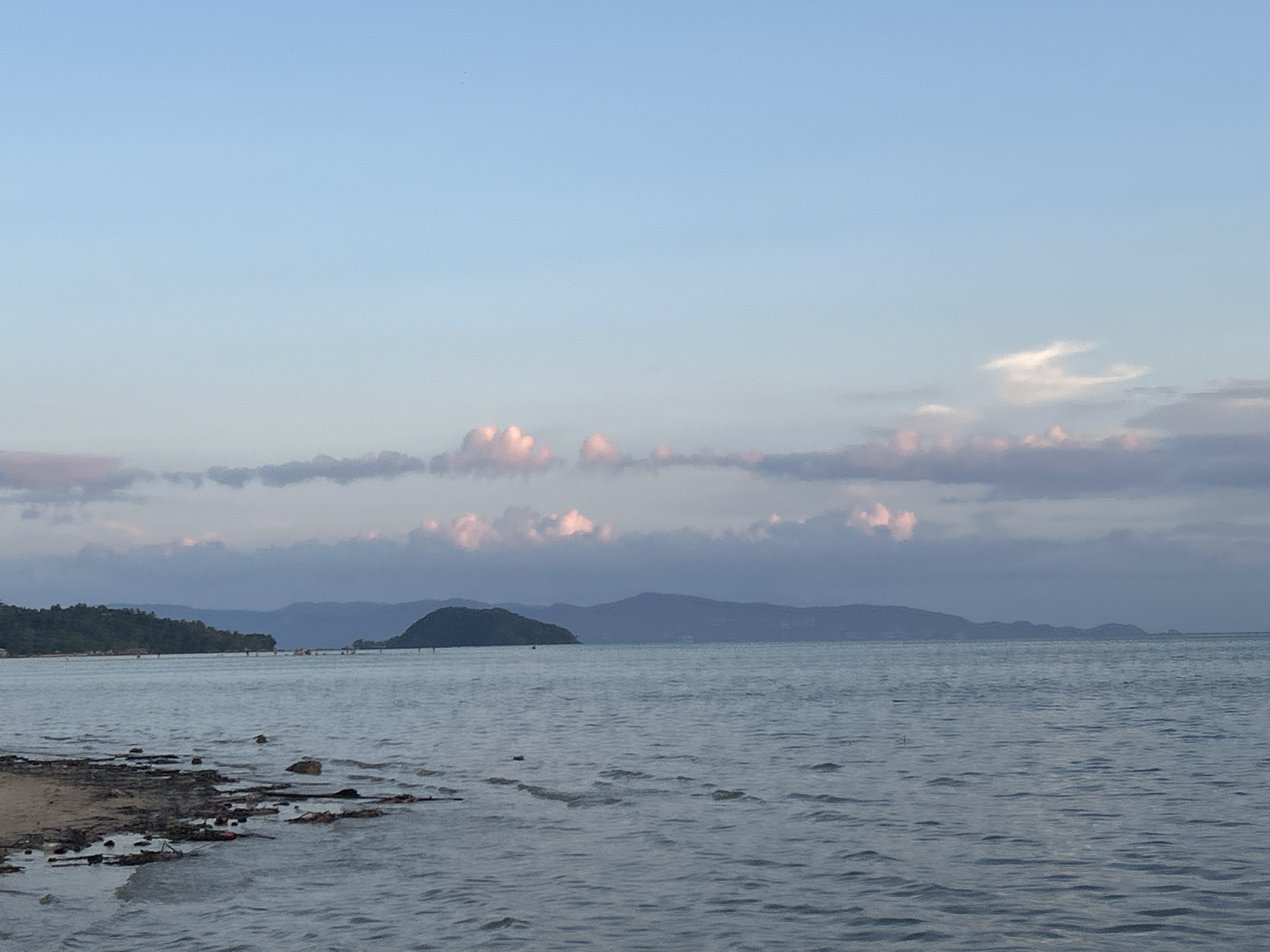 This is a landscape photograph taken in ตำบลเกาะพะงัน, Thailand. The image shows a body of water, likely the sea, with gentle ripples on the surface. In the foreground, there's a shoreline with dark rocks and some sand. Across the water, there is an island or landmass covered in green vegetation. Further in the distance, there are faint mountain silhouettes against the sky. The sky is light blue and filled with clouds, some of which have a pinkish hue, suggesting it might be dusk or dawn.