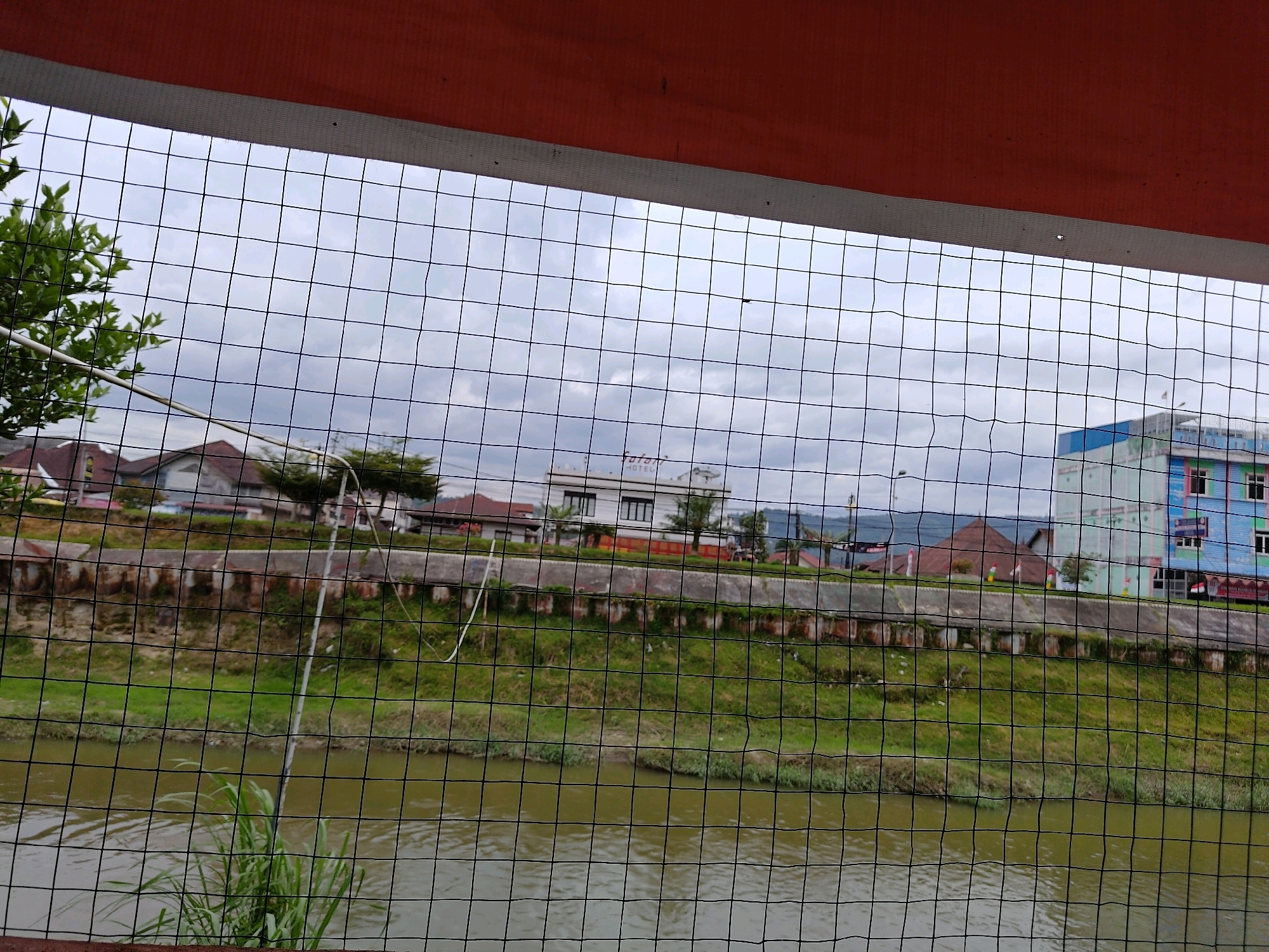 A murky, brown river flows through a city in Tarutung, Indonesia, seen through a black wire fence. The riverbanks are lined with green grass and some shrubbery, with a concrete retaining wall running along the top. Beyond the wall, several buildings are visible, including a white building with "HOTEL" written on it, a red-roofed house, and a brightly colored multi-story building. The sky is overcast with gray clouds. The foreground shows the tops of some green plants growing from the river. The image appears to be taken from a slightly elevated position, possibly a balcony or a bridge.