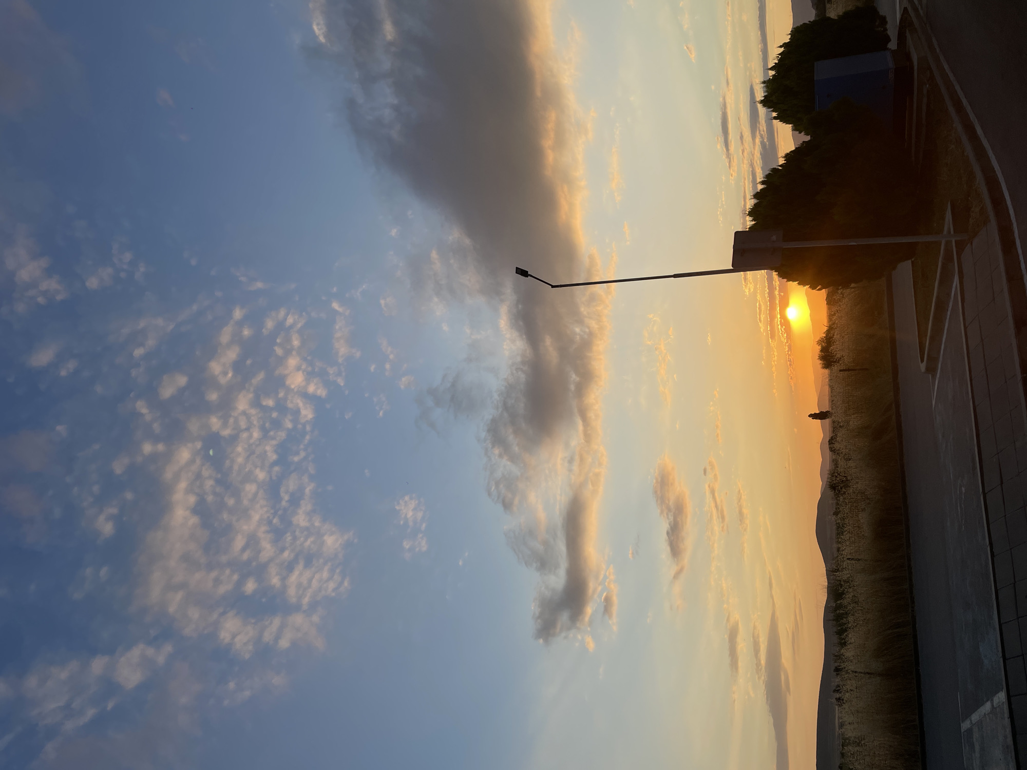 A scenic sunset is captured in Hadzhidimitrovo, Bulgaria. The sky is a blend of blues and oranges, with scattered clouds tinged with pink and yellow. The sun, a bright orb, is low on the horizon, casting a warm glow over the landscape. In the foreground, a paved area, possibly a road or parking lot, is visible. To the right, there are several evergreen bushes and a blue structure, possibly a small building or utility box. A lamppost stands tall near the bushes. Beyond the foreground, a field of dry grass extends towards rolling hills in the distance, silhouetted against the vibrant sunset. The overall atmosphere is peaceful and serene, showcasing the natural beauty of the Bulgarian countryside at dusk.