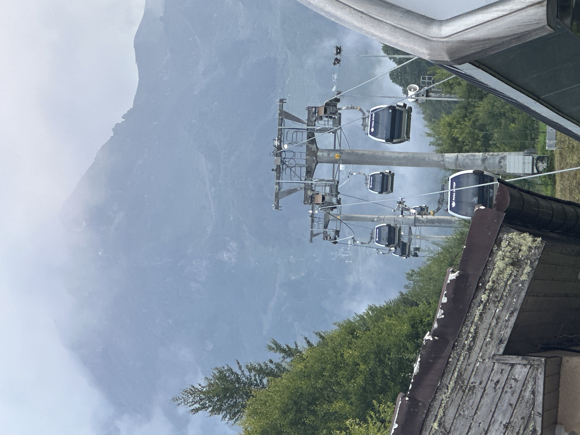 The image depicts a cloudy day in Estosadok, Russia, with a ski lift prominently featured in the foreground and midground. Several cable cars, identified by the Cyrillic text "РОЗА ХУТОР" (Rosa Khutor) visible on their exteriors, are suspended from cables, moving upwards and downwards. These cable cars are modern, black and white, and appear to be carrying passengers.
In the foreground, the weathered wooden roof of a building is visible, showing signs of age with peeling paint and lichen growth. Behind the ski lift, lush green trees cover a hillside, transitioning into the misty slopes of a large mountain range in the background. The mountain peaks are obscured by clouds, creating a sense of depth and atmospheric haze. The overall scene suggests a mountainous resort area, likely operating either in the summer or off-season for skiing.