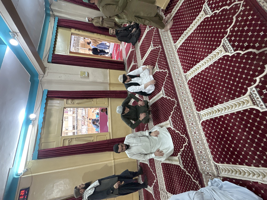 The image displays the interior of a prayer hall, likely a mosque, situated in Quetta City Tehsil, Pakistan. The floor is covered with a deep red carpet featuring a repeating pattern of white and gold prayer arches and borders. The walls are light-colored, with a pale blue trim along the ceiling and above window/door frames. Fluorescent light fixtures are visible on the ceiling. A digital clock on the upper left wall displays "3:23". Red curtains frame multiple rectangular openings.
Through a window on the mid-left, an exterior urban environment with multi-story buildings and several individuals is visible, with a pink/purple structure or surface present below the window line. A doorway on the mid-right opens to an outdoor, possibly covered, area where additional men are standing or moving.
Approximately ten to fifteen adult males are present within the scene, predominantly dressed in traditional South Asian attire such as shalwar kameez. In the foreground, five men are engaged in prayer or prayer-related postures. From left to right:
1. A man standing on the far left, wearing a dark shalwar kameez and a light checkered shawl over his shoulder and head, observing the scene.
2. A man kneeling on a prayer mat, facing away from the camera, wearing a light-colored shalwar kameez and a dark beard, in a sitting posture.
3. A man crouching on a prayer mat, wearing a dark green shalwar kameez and a light cap, with hands resting on his knees.
4. A man sitting cross-legged on a prayer mat, wearing a white shalwar kameez, a dark waistcoat, and a light cap.
5. A man partially visible on the far right, in an olive-green shalwar kameez, standing barefoot.
Further back, a man in a dark outfit is seated on the carpet, and several other men are visible through the open doorway and in the exterior view through the window, some standing, others gathered around an unseen object.