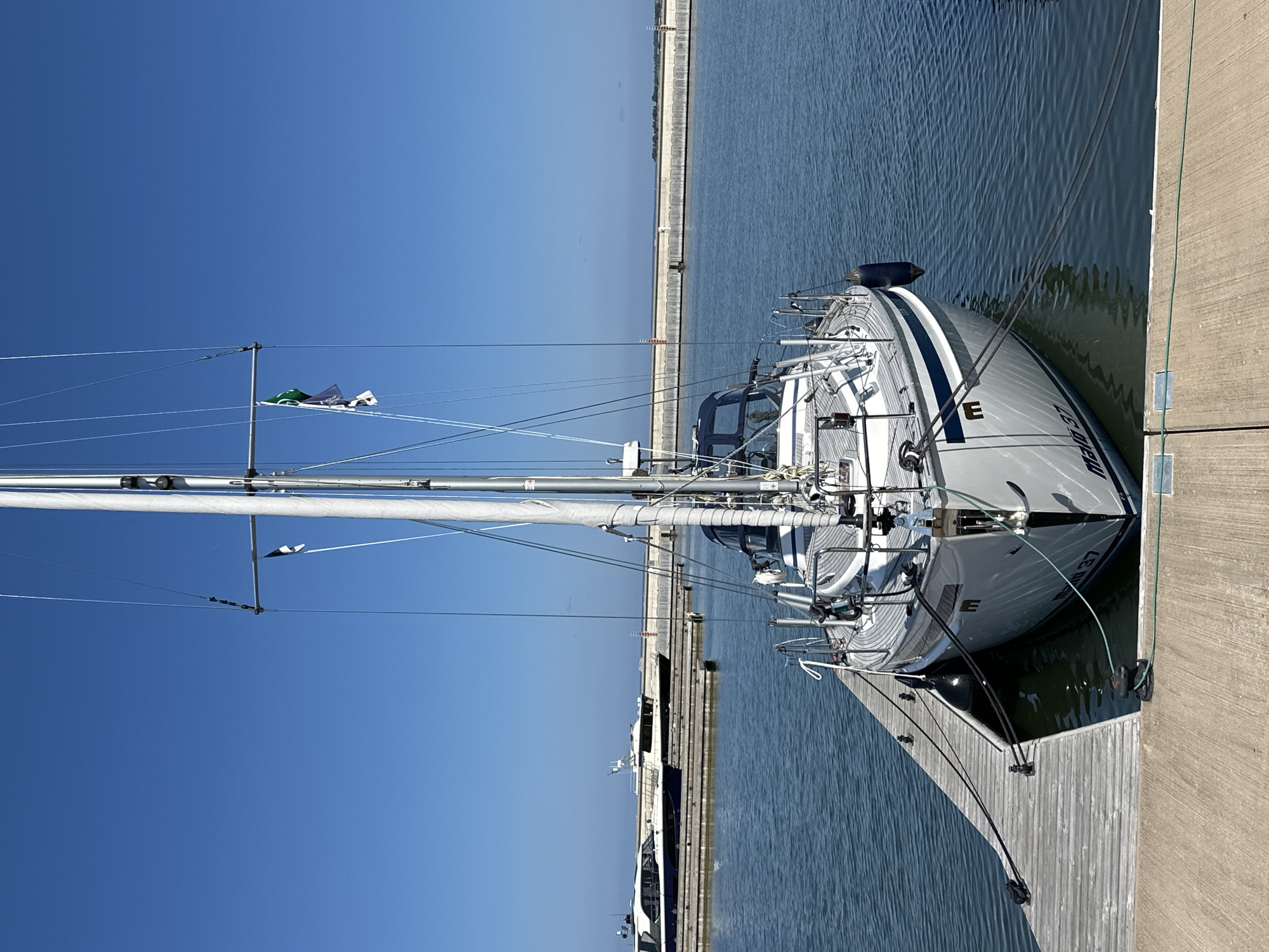 A modern white sailboat named "Mala 37" is docked at a wooden pier on a clear, sunny day in Tallinn, Estonia. The boat is positioned in the center of the frame, facing the viewer. Its mast is tall and slender, with rigging and several flags attached. The hull is white with a blue stripe. The pier is made of weathered grey planks and is secured by black ropes to the boat. In the background, other boats and a long concrete breakwater are visible across the calm blue water. The sky is a vibrant, cloudless blue. The overall scene depicts a peaceful harbor setting.