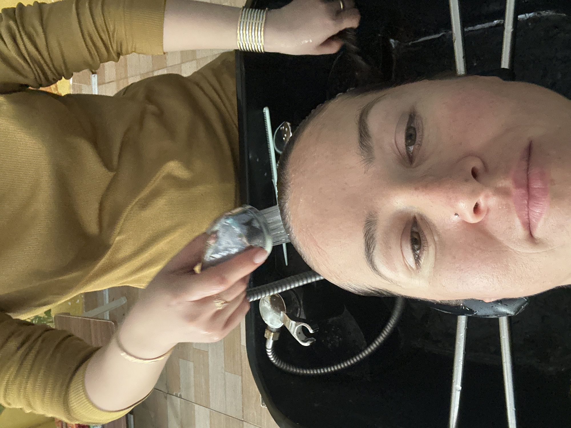 A woman lies with her head back in a black salon hair washing sink. A person wearing a long-sleeved, mustard-colored shirt is holding a shower head and rinsing the woman's hair. The shower head is silver with a clear plastic attachment. The woman has fair skin, brown eyes, and light eyebrows. She has a stud piercing on the left side of her nose. She is wearing lipstick. The person rinsing her hair has gold bangles on their wrist and a gold ring on one of their fingers. The sink has a silver shower hose and faucet. A comb is visible on the left side of the sink. The background includes tiled flooring. The location is likely a hair salon, but no specific location in Bac Nha Trang Ward, Vietnam can be confirmed.
