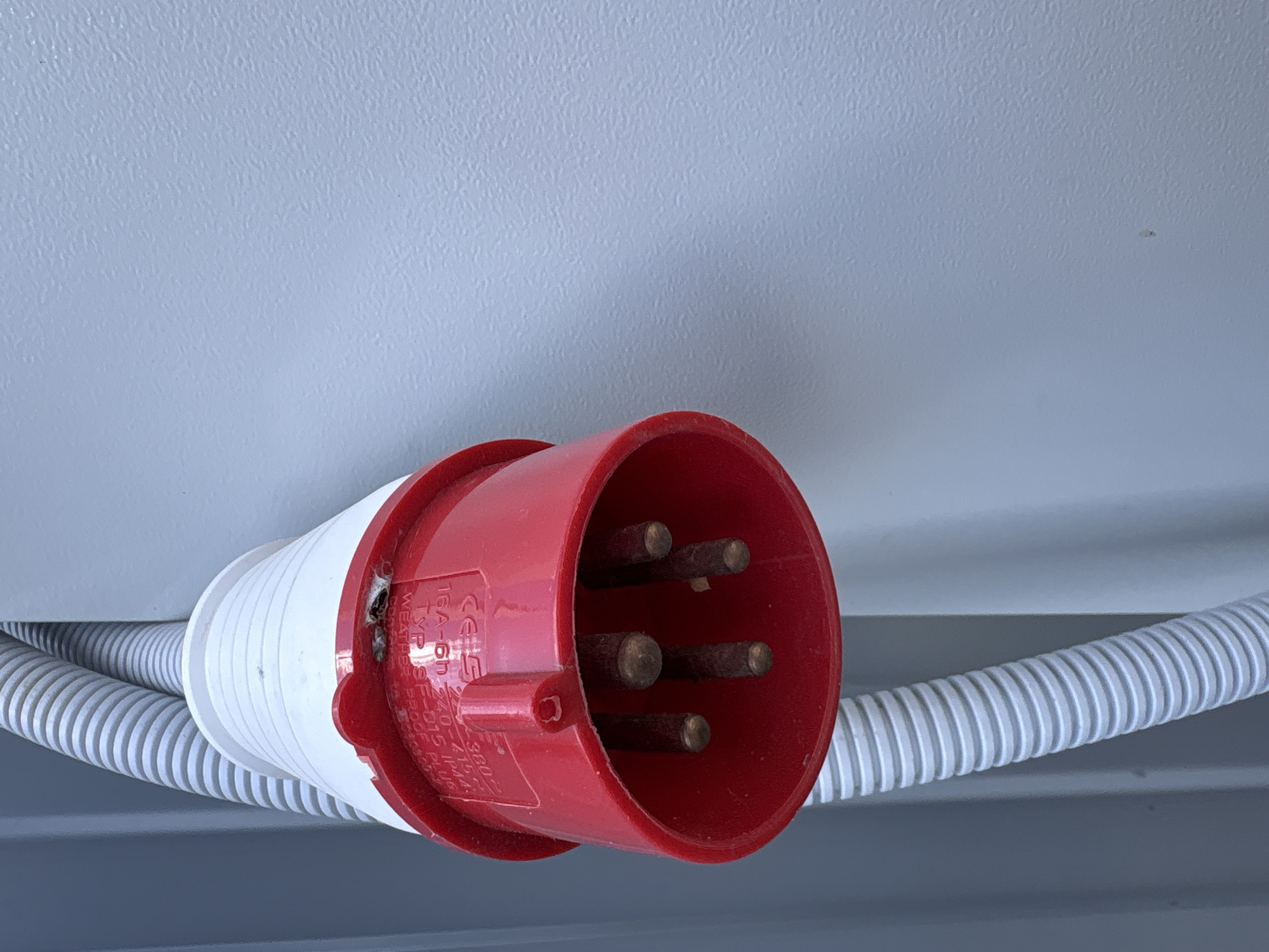 A close-up shows a red and white industrial electrical plug with five pins, connected to a white corrugated conduit. The plug is mounted on a light gray surface. The location is not discernible from the image.