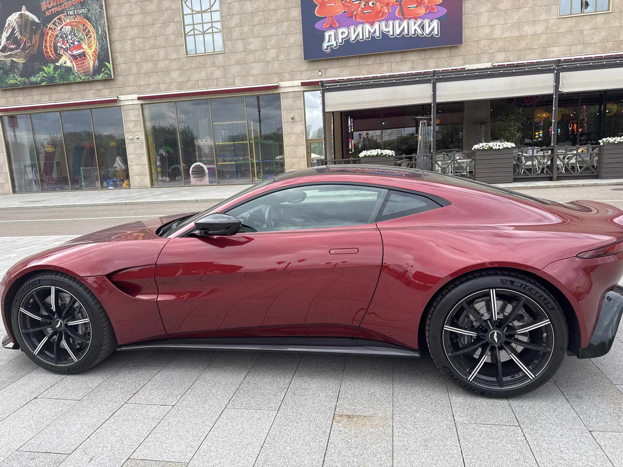 A red Aston Martin Vantage is parked on a gray tiled pedestrian walkway in front of a modern building. The building has large windows, awnings, and outdoor seating areas for a restaurant. Above the awnings, large advertisements are visible: one features a dinosaur and a roller coaster, and another has cartoon characters with the Russian word "ДРИМЧИКИ" (Dreamchiki). The car is viewed from the side, showcasing its sleek design and black alloy wheels with white accents. The environment appears to be a public area, possibly a shopping center or entertainment complex. There are no people visible in the image. The lighting suggests it is daytime.